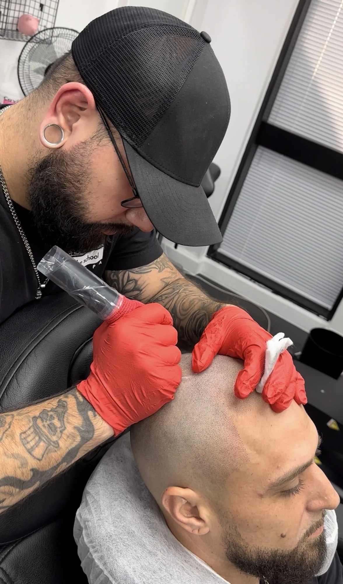 Tattooed man receiving a tattoo on his head from a tattoo artist wearing red gloves in a tattoo studio.