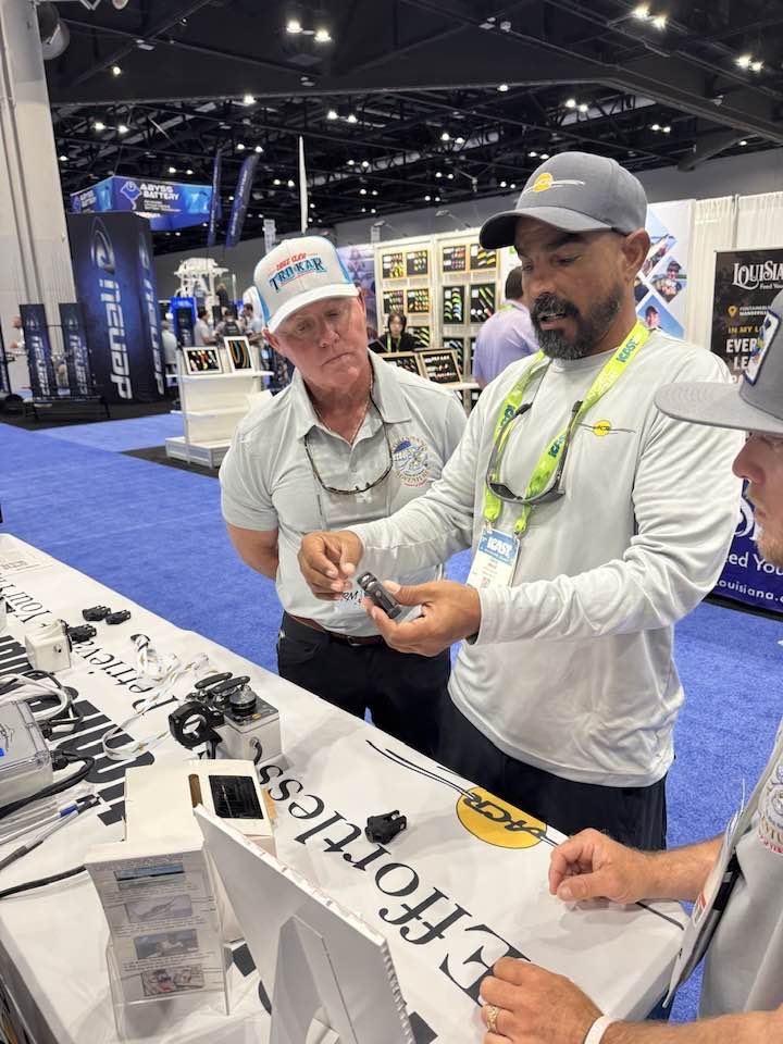 Three men inspecting fishing or boating gear at an indoor trade show, with displays and booths in the background.