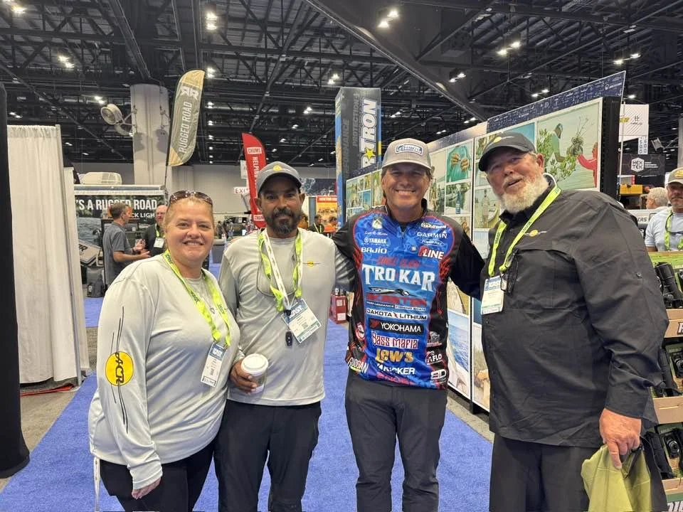 Four people smiling and posing at a trade show or convention booth, with banners and display boards in the background.