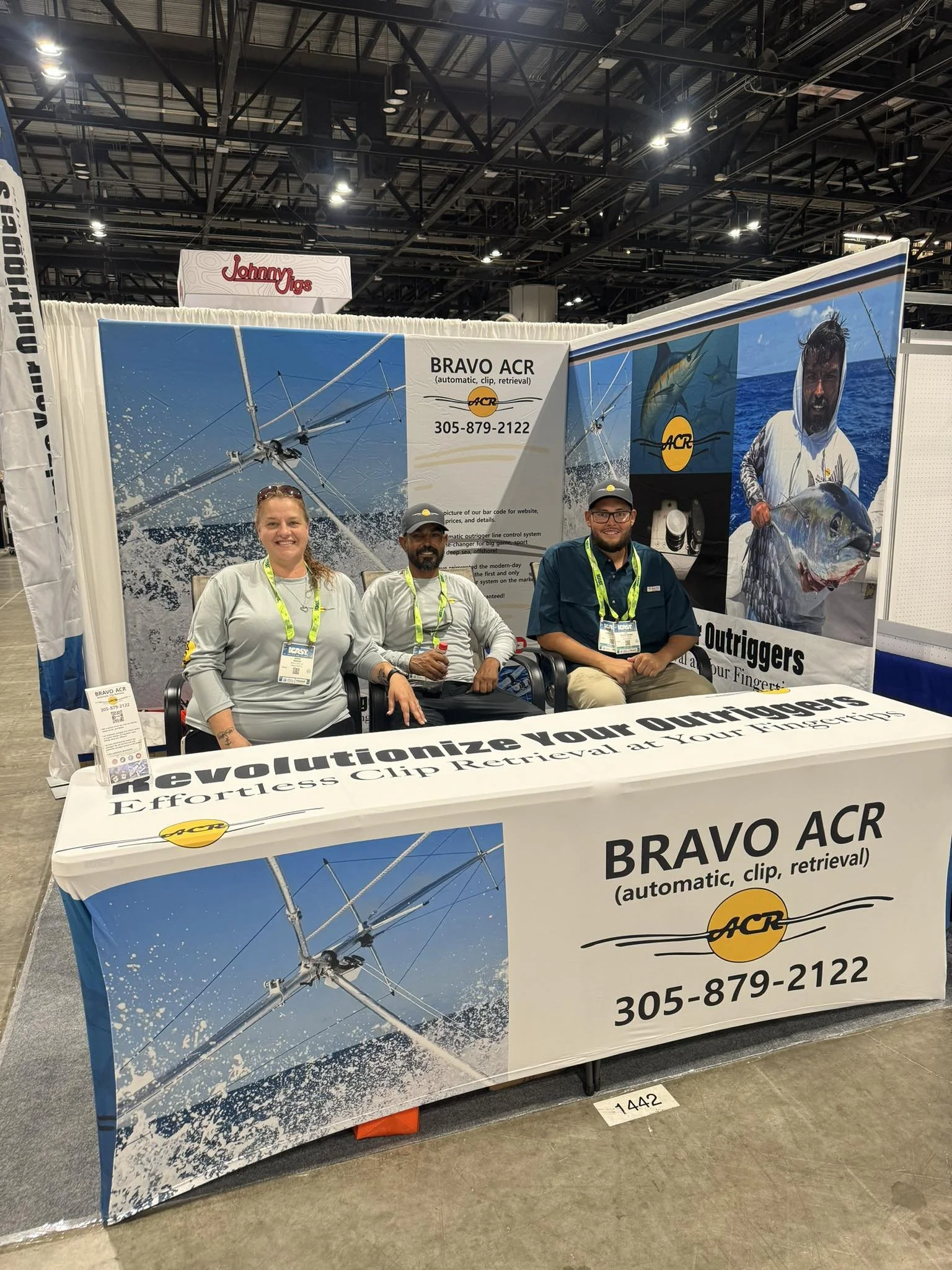 Three people sitting at a booth promoting Bravo ACR, a company that offers automatic, clip, and retrieval systems for fishing outshooters. The booth features banners with images of fishing equipment and a man holding a large fish. The tablecloth displays the company's name, contact number, and slogan, "Revolutionize your Outshooters, Effortless Clip Retrieval at Your Fingertips." The setting appears to be an indoor trade show or expo.