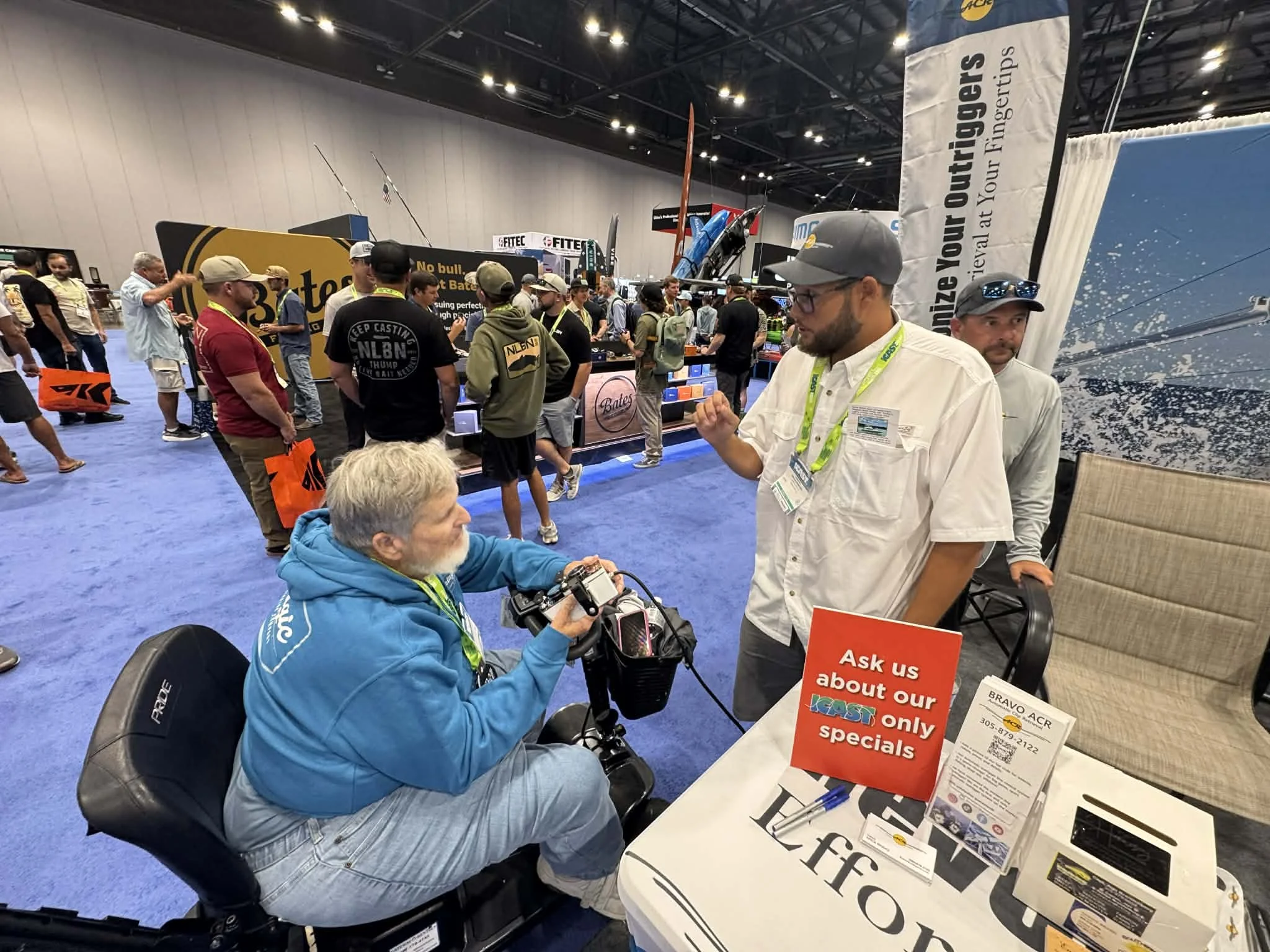 At a trade show, a man with gray hair and beard in a blue hoodie is seated in a mobility assist device, holding a device and talking to a man with a cap and glasses, who is standing. There is a sign on the table that says 'Ask us about our FAST only specials'.