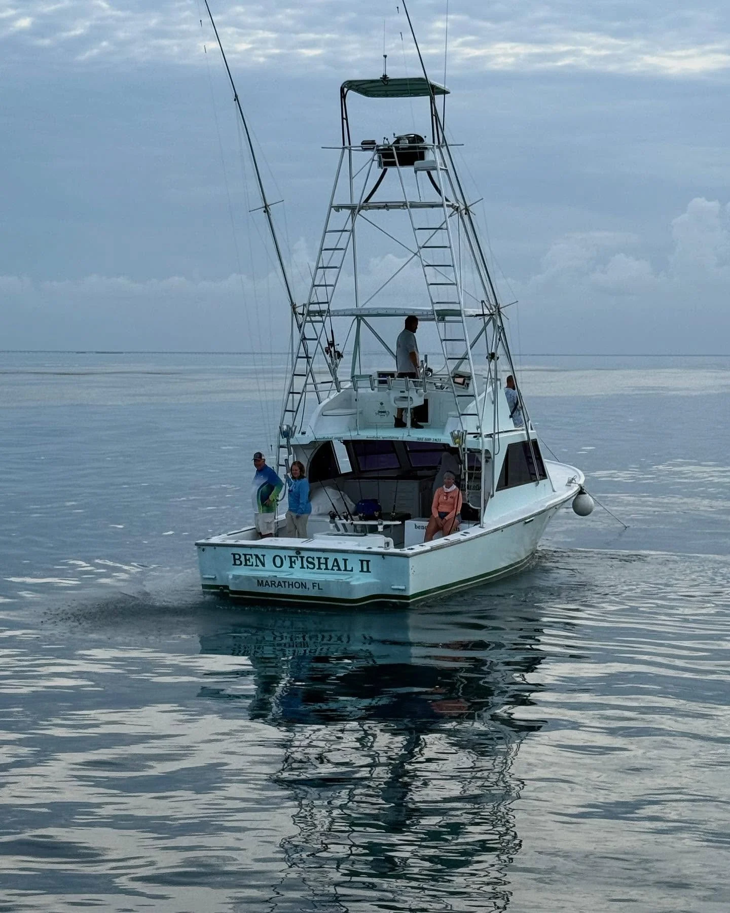 A fishing boat named Ben O'fishal II from Marathon, FL, on calm water with four people onboard, under a cloudy sky, partial view of water reflection.