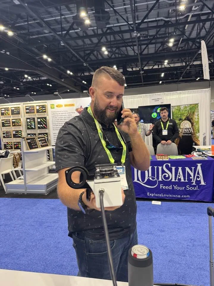 A man with a beard and short hair, wearing a black shirt, is standing at a booth holding a white device with a strap, talking on a phone. There is a can of drink in front of him. The background has a booth display with a Louisiana banner that reads 'Louisiana Feed Your Soul,' and has posters, a TV, and other people.