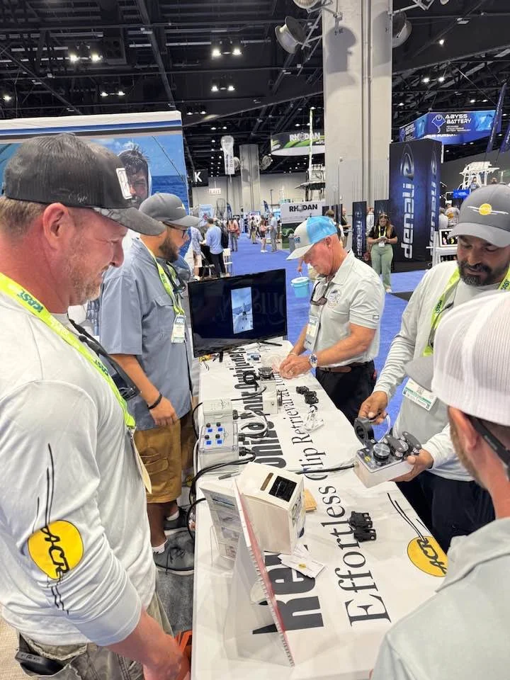 A group of men at an exhibition booth are demonstrating remote control devices for boats or ships. They are surrounded by various electronic equipment, a monitor, and promotional material with a banner that reads "Elite" and "Effortless".