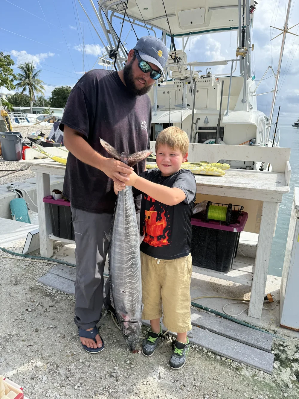 A man and a boy holding a large fish on a dock with boats in the background.