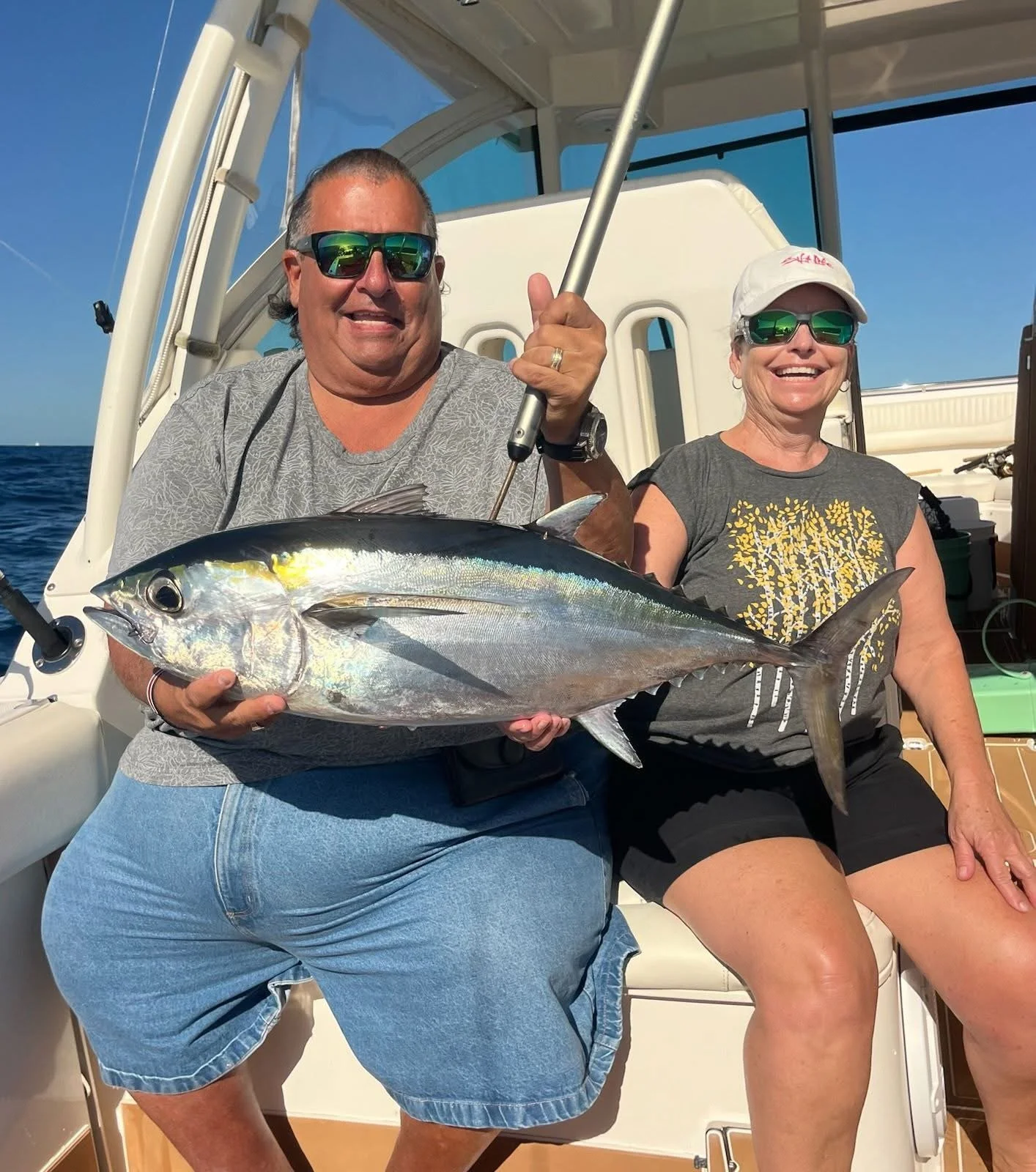 Two smiling people on a boat holding a large fish, with a clear blue sky and ocean in the background.