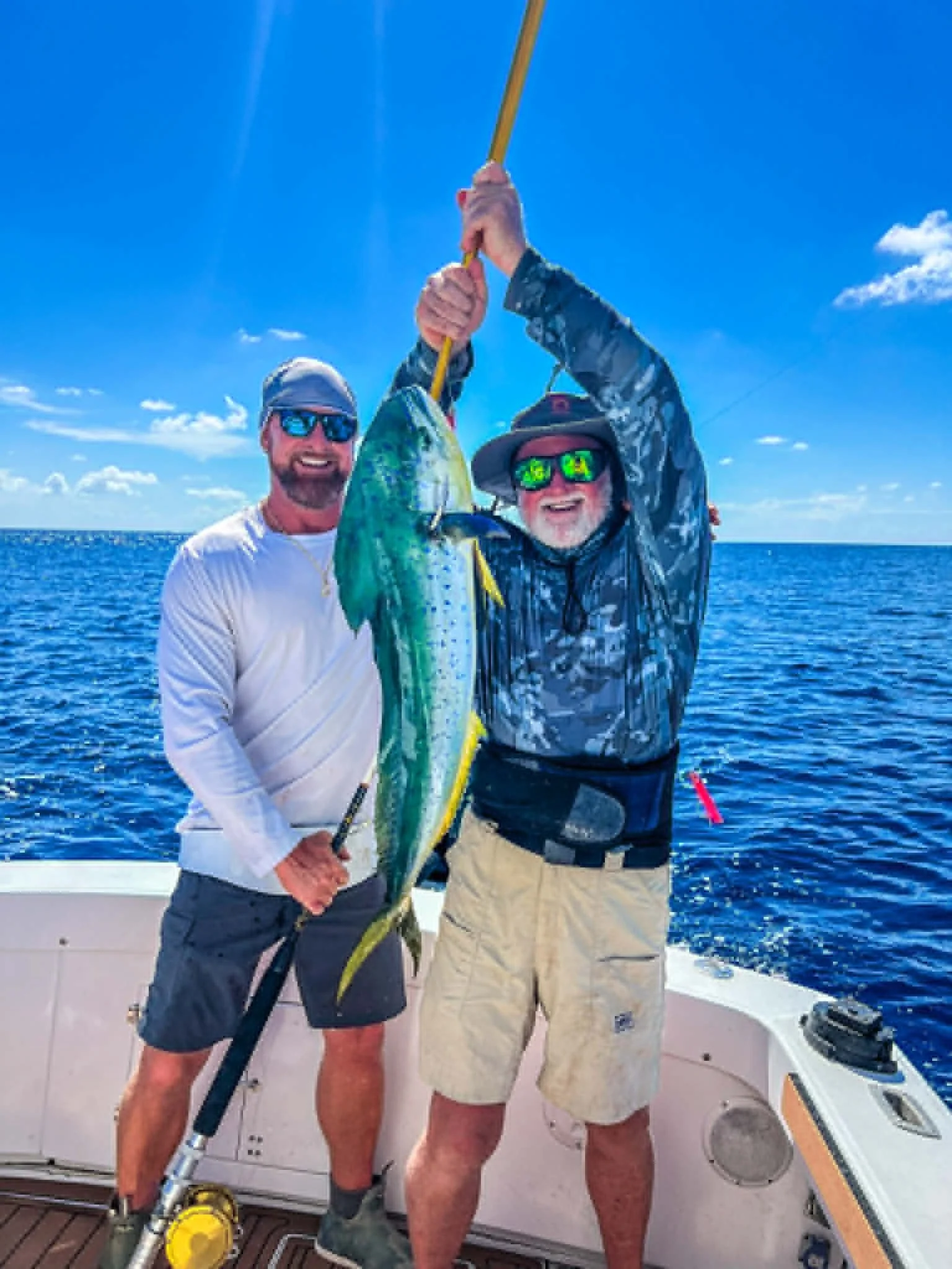 Two men on a boat at sea holding a large fish. They are smiling, wearing sunglasses and hats, with a clear blue sky and ocean in the background.
