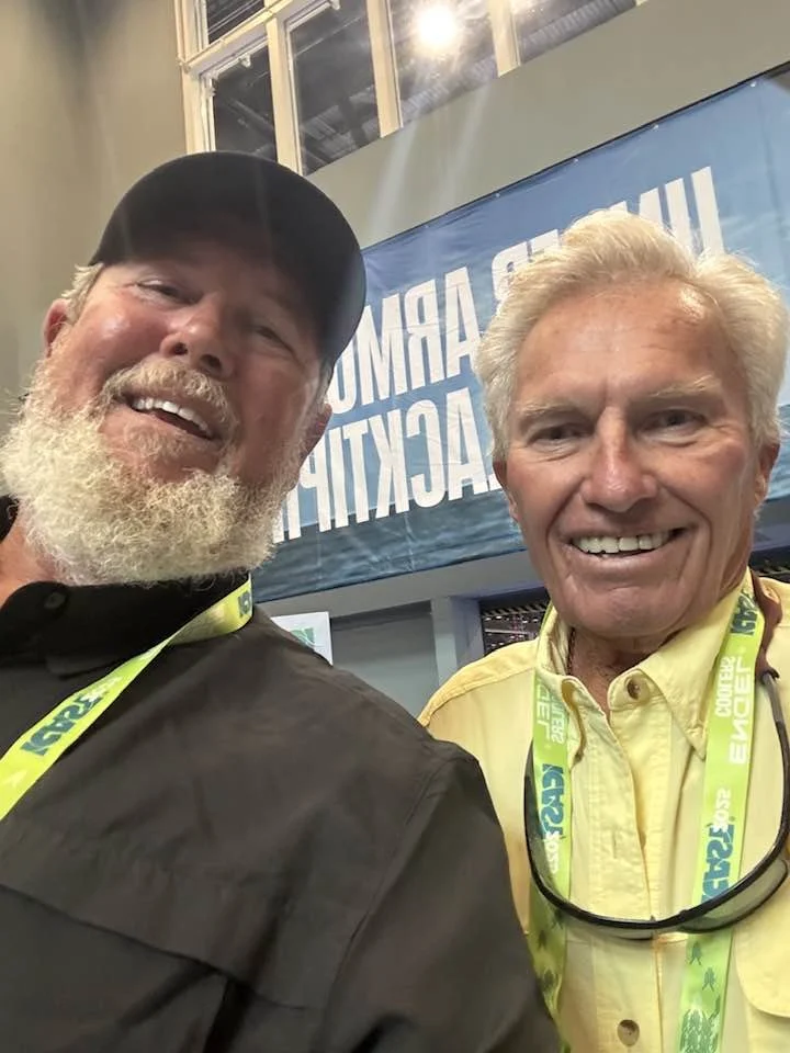 Two elderly men smiling for a selfie at an indoor event with a blue banner in the background that has white text, and they are wearing event lanyards around their necks.