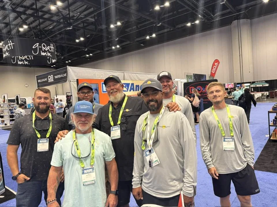 A group of seven men standing together at a trade show, smiling, wearing lanyards and badges. The background features booths, banners, and other attendees in an indoor exhibition hall.