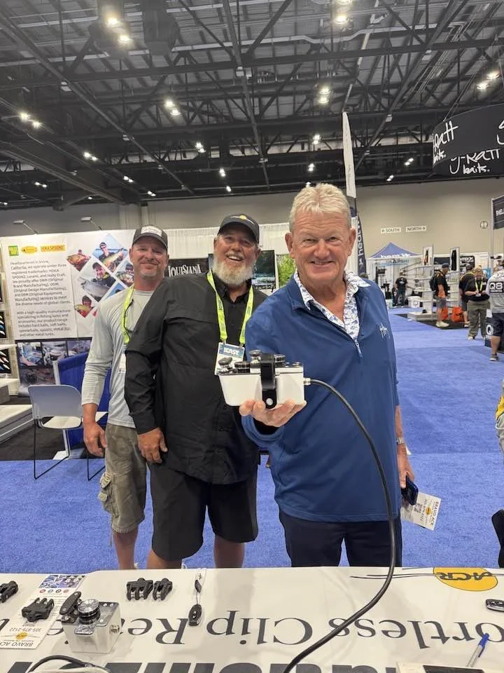 Three men standing inside a convention center, smiling at the camera. The man in front is holding a device with a cable attached. The background features display booths and banners.