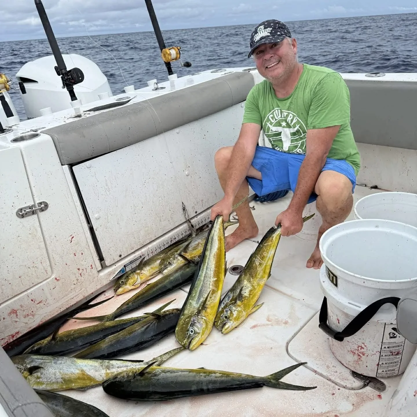 A man kneeling on a boat holding two large fish, with several other fish laid out on the deck, at sea during daytime.