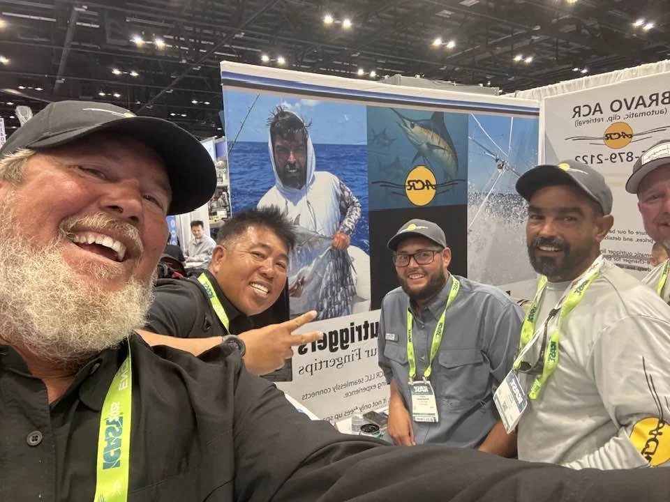 Five men at a fishing industry trade show, smiling for a selfie in front of a booth with fishing-related imagery and logos, wearing conference lanyards.