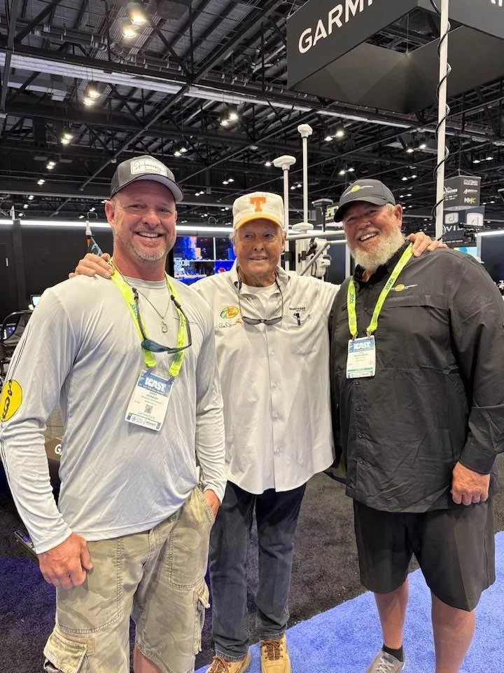 Three men smiling and posing together at an indoor trade show, with a Garmin sign visible in the background. All are wearing casual outdoor clothing, with two of them wearing baseball caps and lanyards with exhibitor badges.