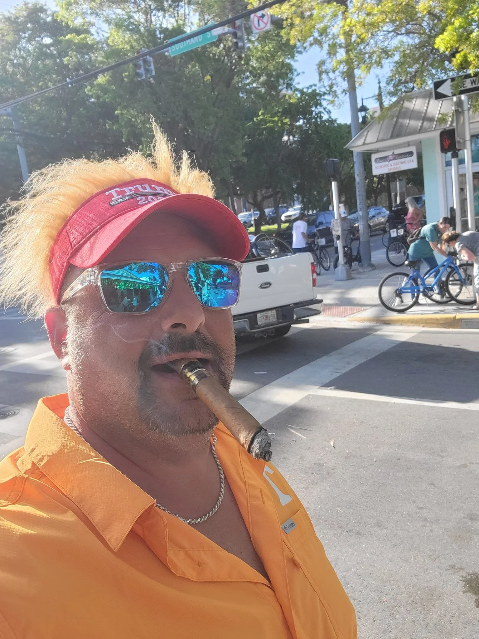A man with blond spiky hair, sunglasses, and a Trump 2020 hat smokes a cigar on a city street. He is wearing an orange shirt and a silver chain necklace. In the background, there are people and bicycles on the sidewalk, a white truck, and traffic signals.