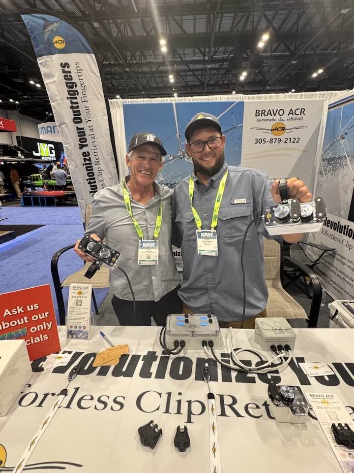 Two smiling people at a trade show booth displaying remote clutch release devices. The woman on the left holds a device, and the man on the right shows a similar device. Both are wearing badges and lanyards.
