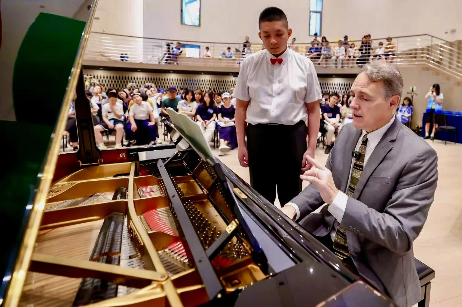 A man in a gray suit and tie teaches a young boy in a white shirt and red bow tie how to play piano in front of an audience in a large room with high ceilings and windows.