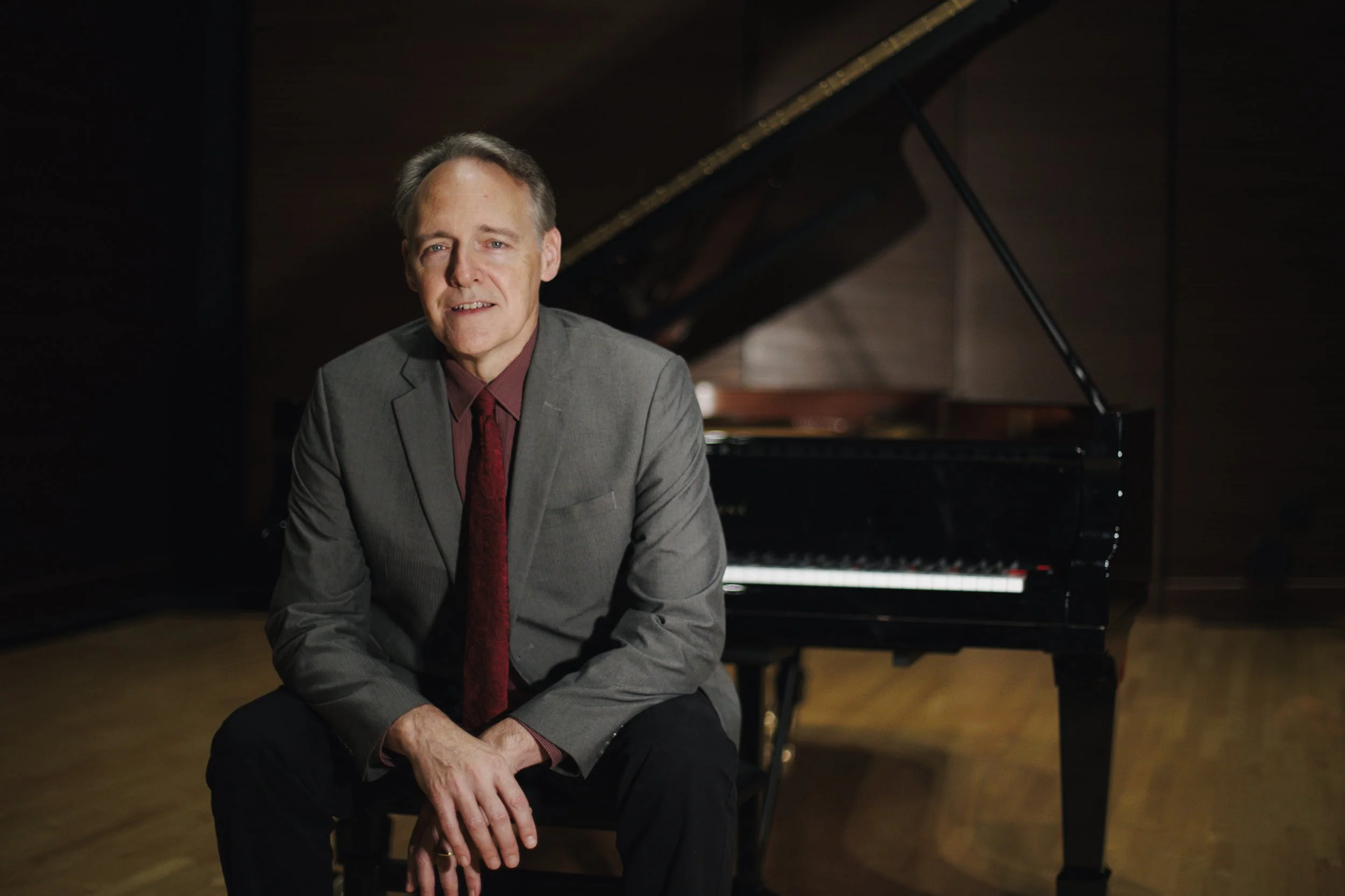 A man sitting next to a grand piano in a dimly lit music studio or concert hall.