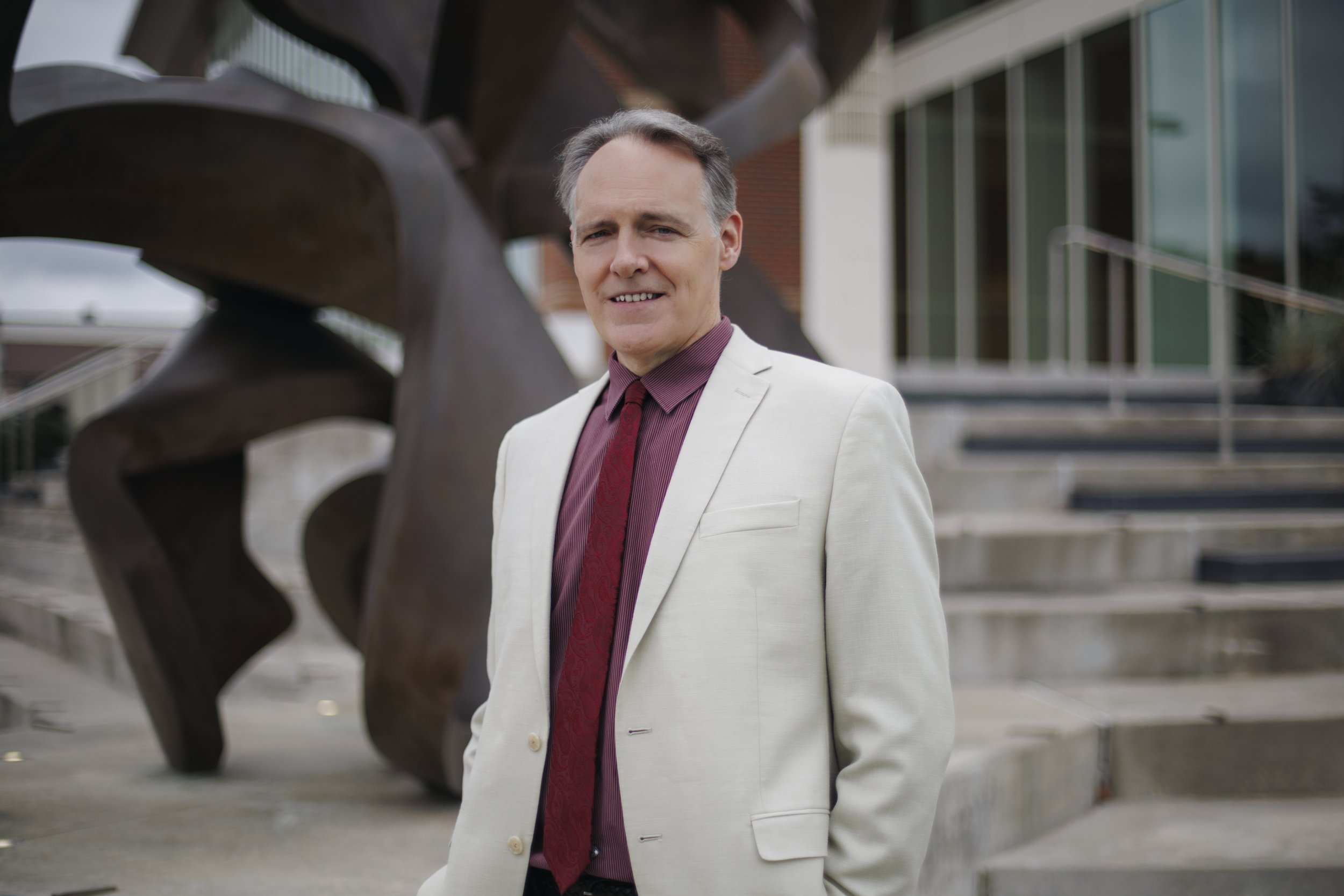 A middle-aged man in a white suit and red shirt with a red tie, standing outdoors in front of metal stairs and an abstract sculpture.