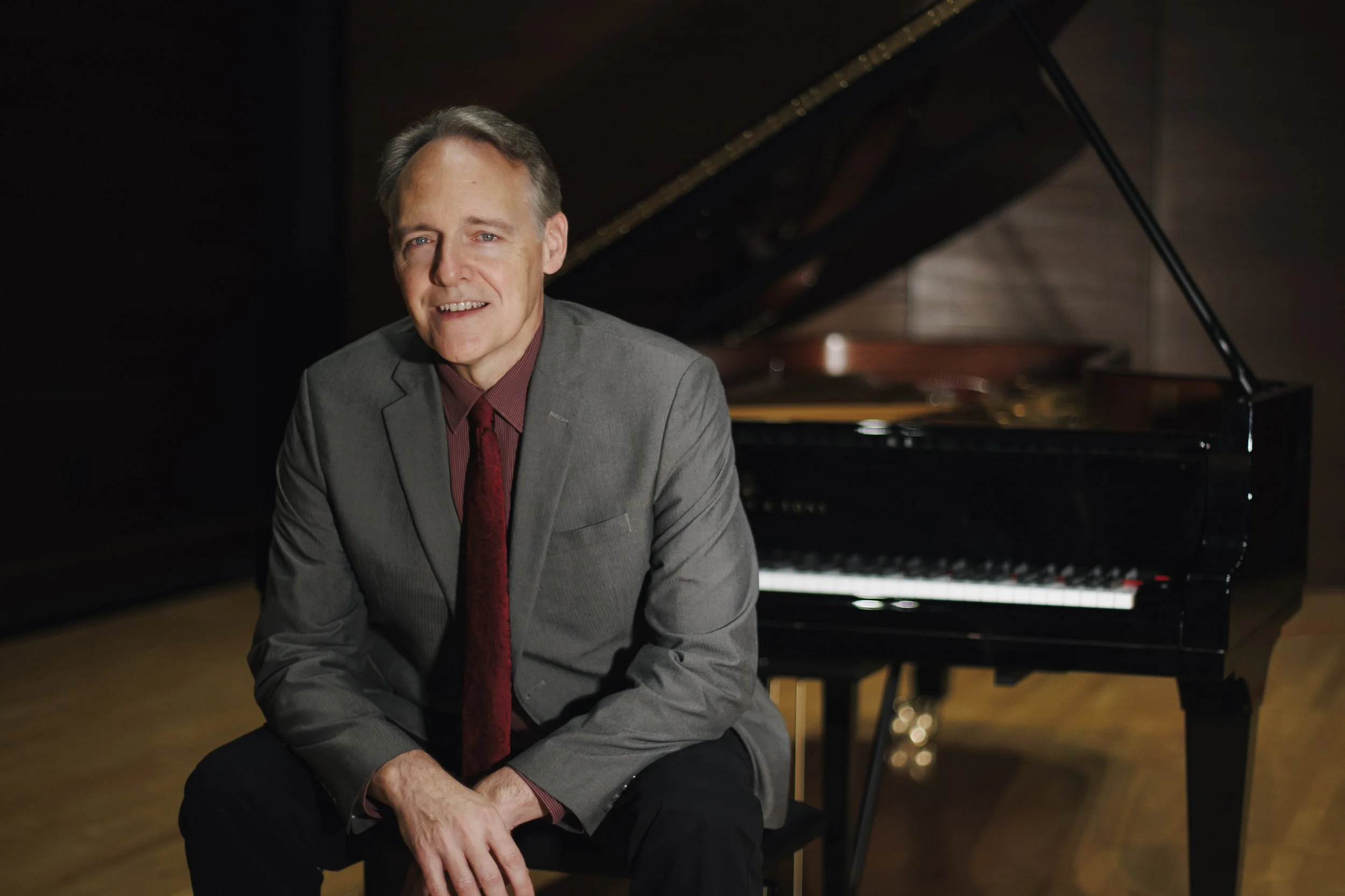 A man in a grey suit and red tie sitting in front of a grand piano in a music studio.