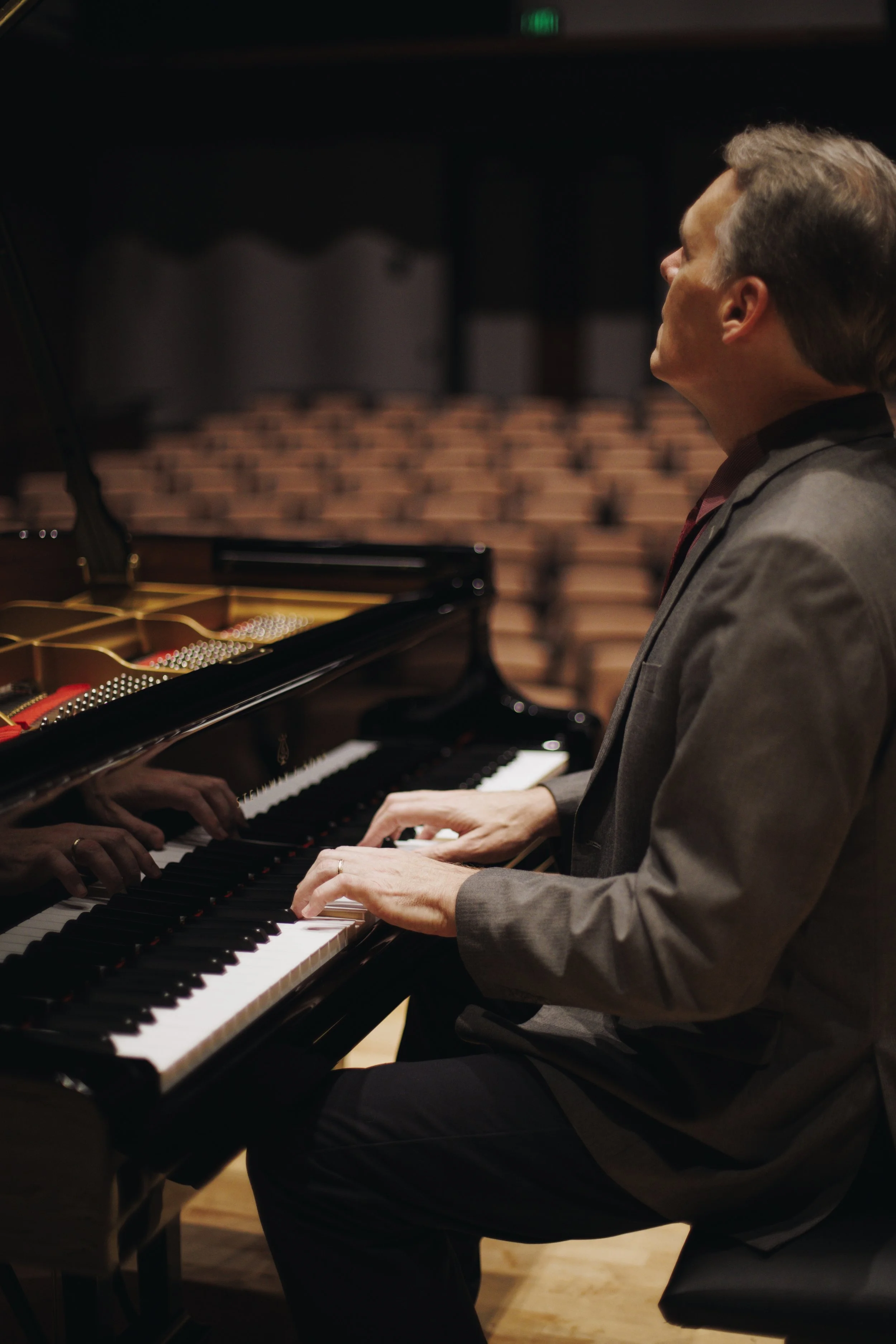 A man in a gray blazer playing a grand piano in a concert hall.