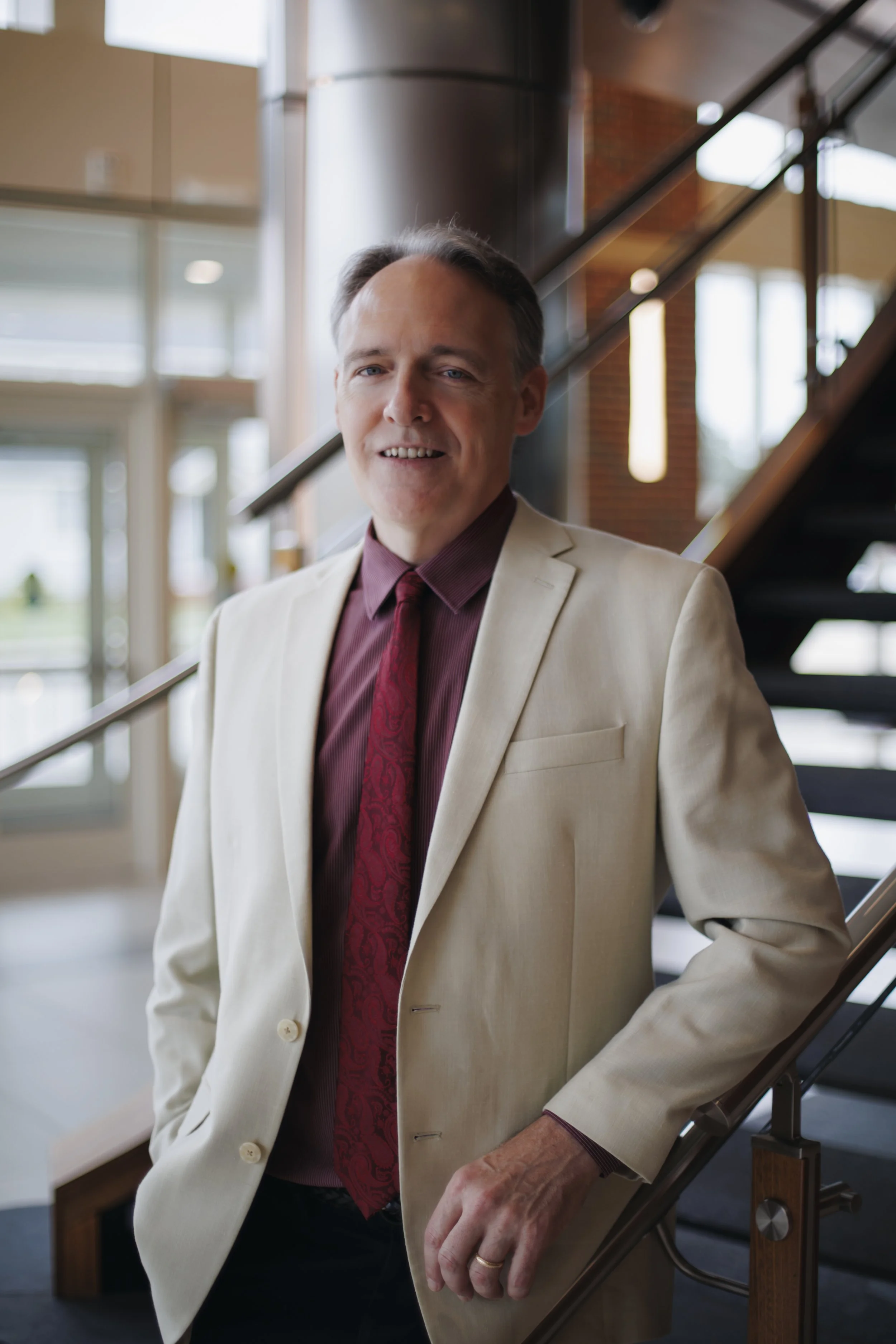 A man wearing a beige suit and a red tie standing on a staircase inside a modern building with large windows.