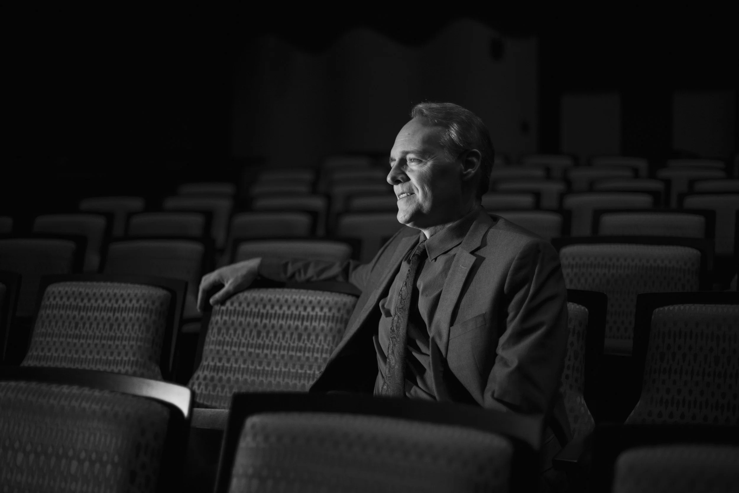 A man in a suit sitting alone in an auditorium with rows of empty seats, illuminated by a spotlight.