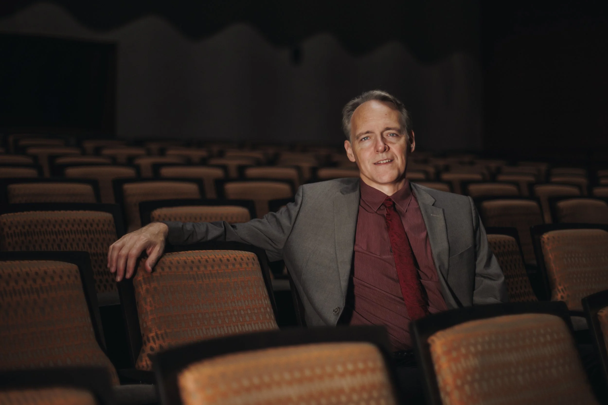 A man in a gray suit and burgundy shirt and tie sitting alone in an empty theater with rows of orange and black seats.