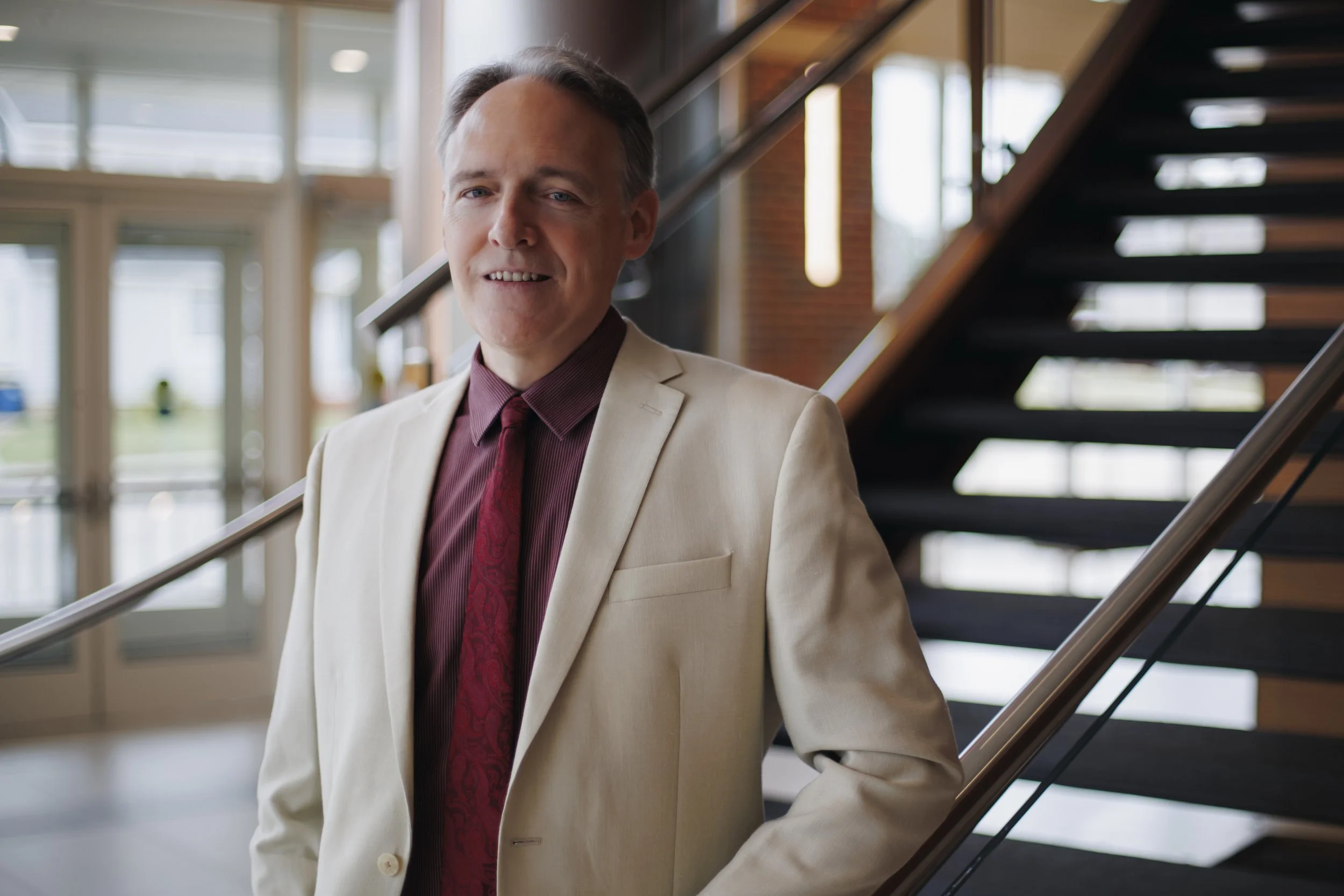 A man with short gray hair, wearing a cream-colored suit jacket, maroon shirt, and red tie, standing indoors near a staircase with glass doors and windows behind him.