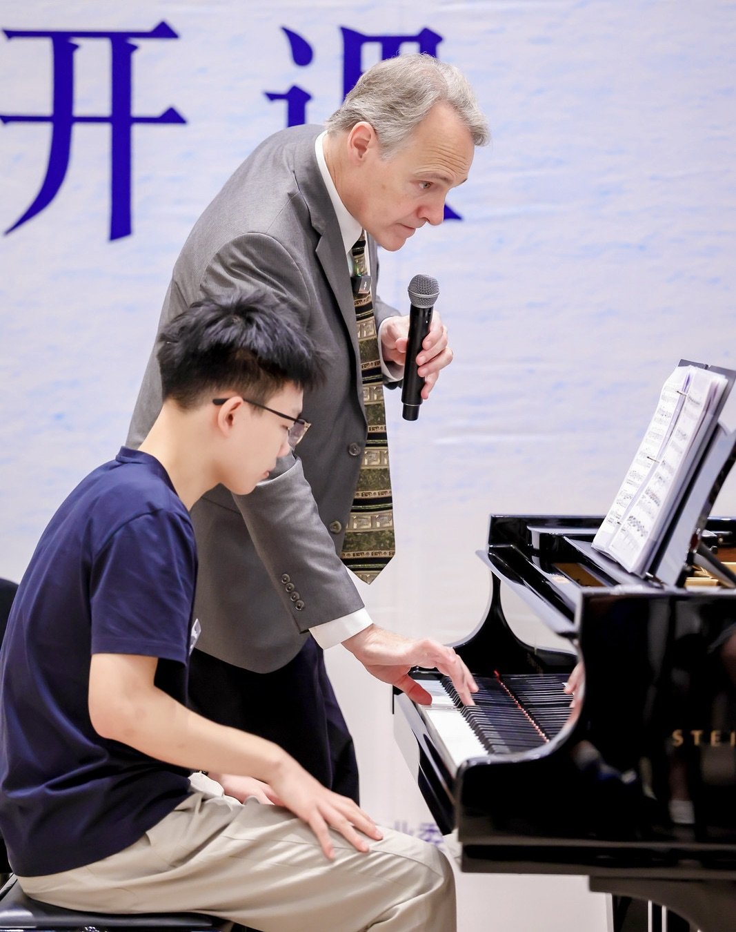 An older man in a suit with a microphone helping a young man with glasses play piano at a music lesson or performance.