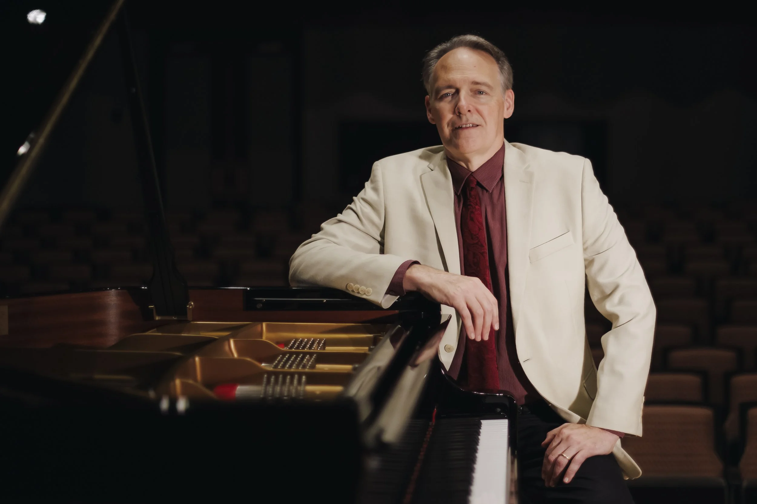 A man in a cream blazer and red shirt sitting at a grand piano in a dimly lit auditorium.