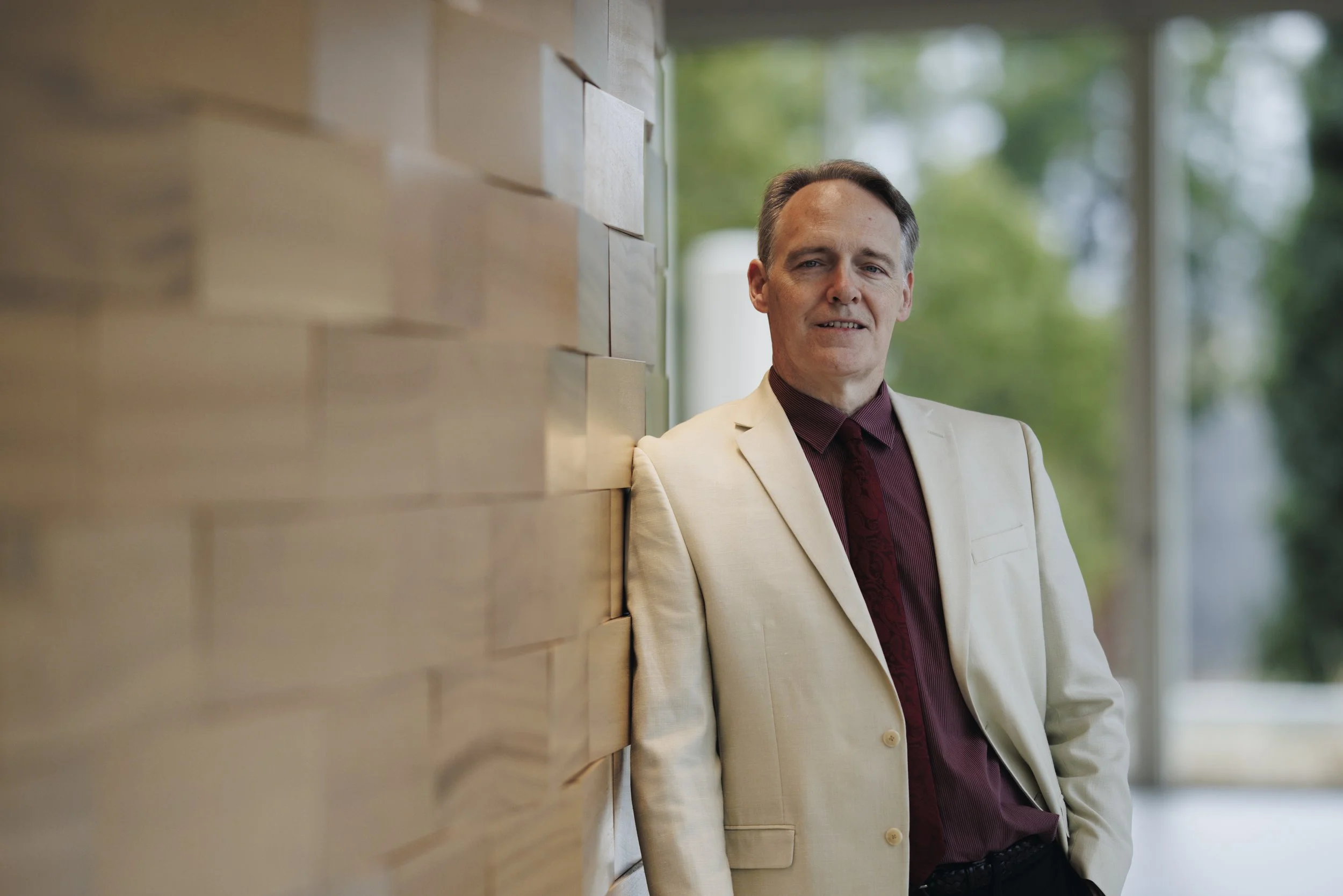 A middle-aged man in a cream-colored suit leaning against a wooden wall, with a background of large windows and trees outside.