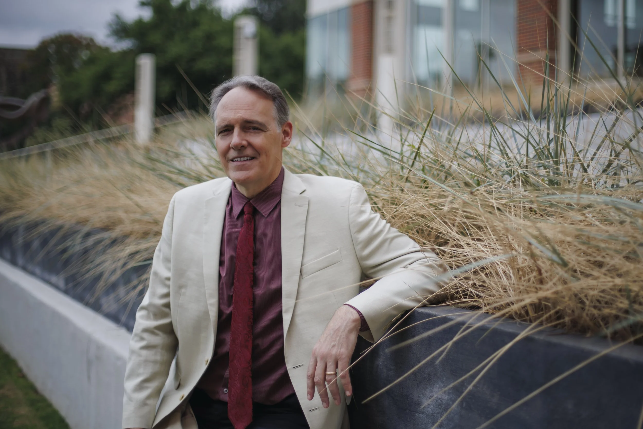 A middle-aged man in a cream-colored suit jacket, maroon shirt, and matching tie, leaning against a black railing, smiling outdoors with tall dry grass and a modern building in the background.