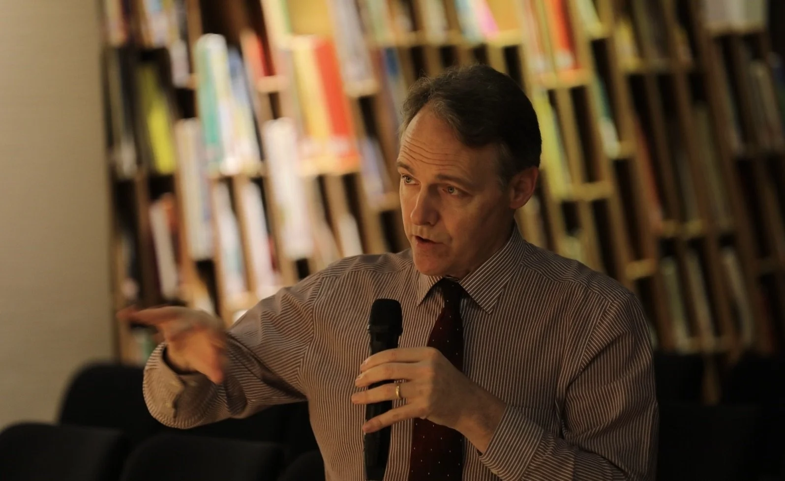 A man in a striped shirt and tie is speaking into a microphone while gesturing with his hand in a room with a colorful bookshelf in the background.