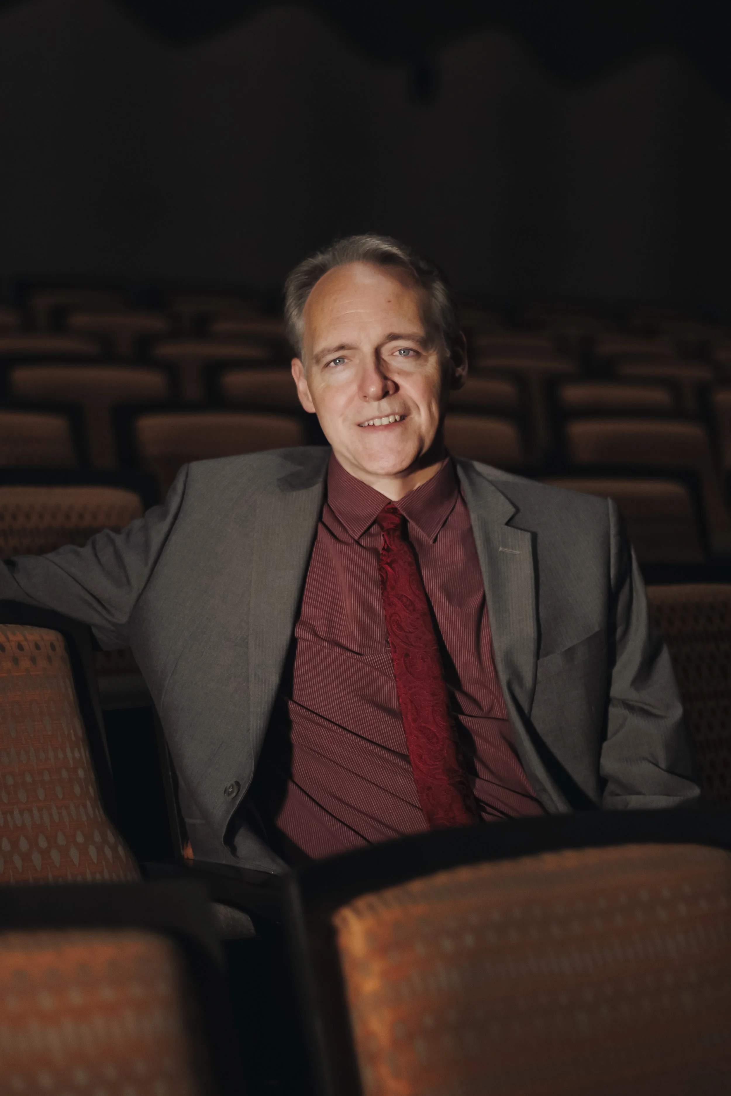 A man in a gray suit and maroon shirt with a patterned tie sitting in an empty theater with multiple rows of brown seats.