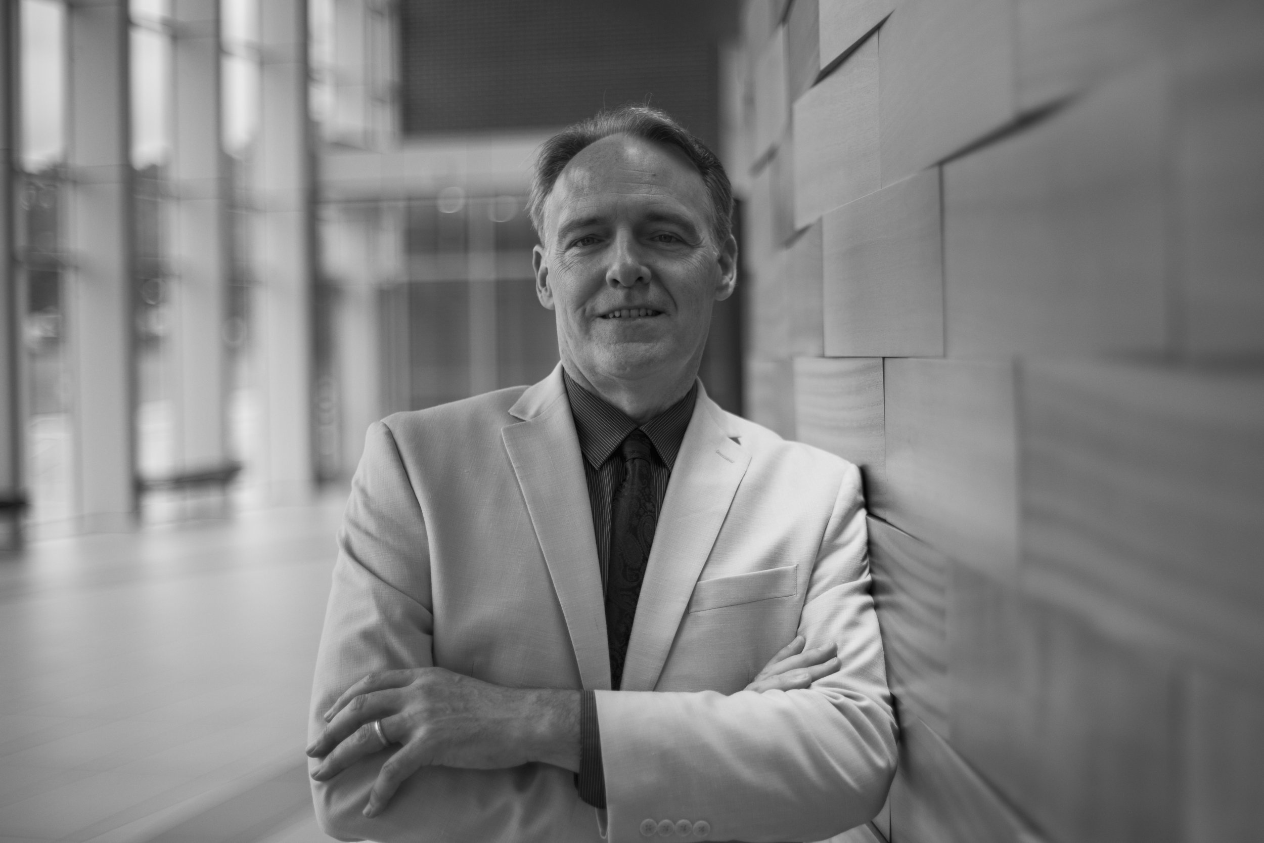 Black and white photo of a man in a light-colored suit leaning against a wall with arms crossed, smiling at the camera in a modern indoor space.