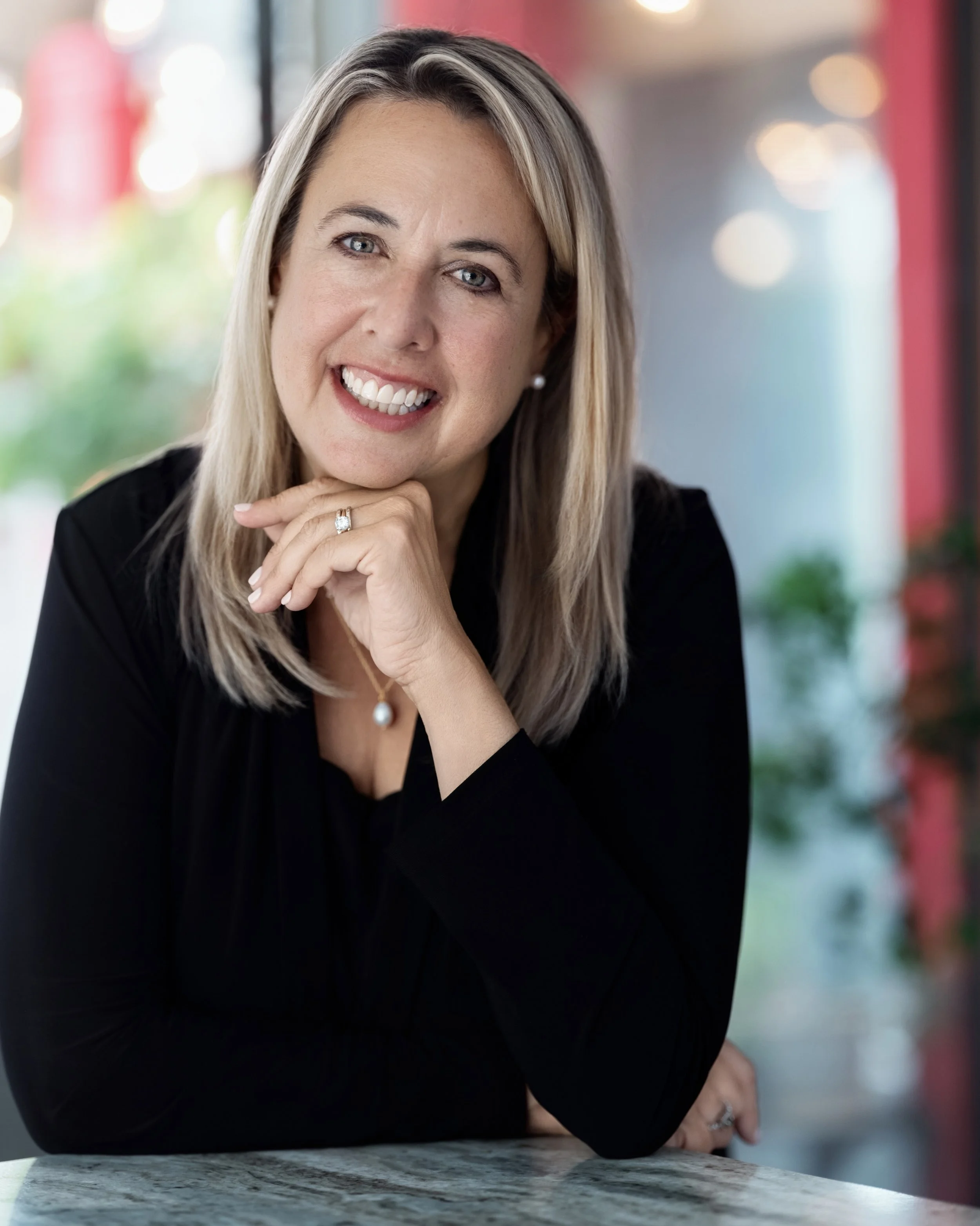 A woman with shoulder-length blonde hair, smiling, resting her chin on her hand, wearing a black top and pearl jewelry, sitting at a marble table with a blurred background of colorful lights and greenery.