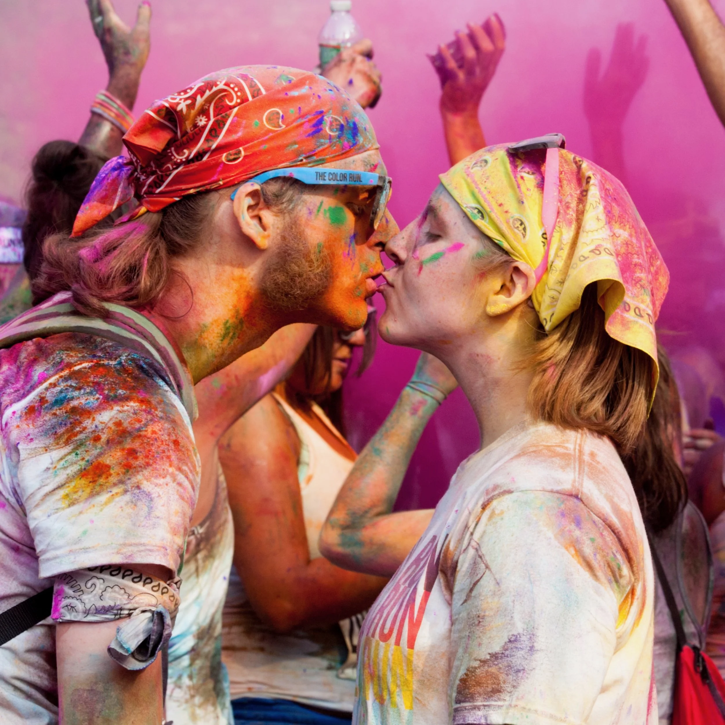 A man and a woman are about to kiss, both covered in colorful powder from a color run event, wearing bandanas on their heads, with other participants in the background also covered in colors.
