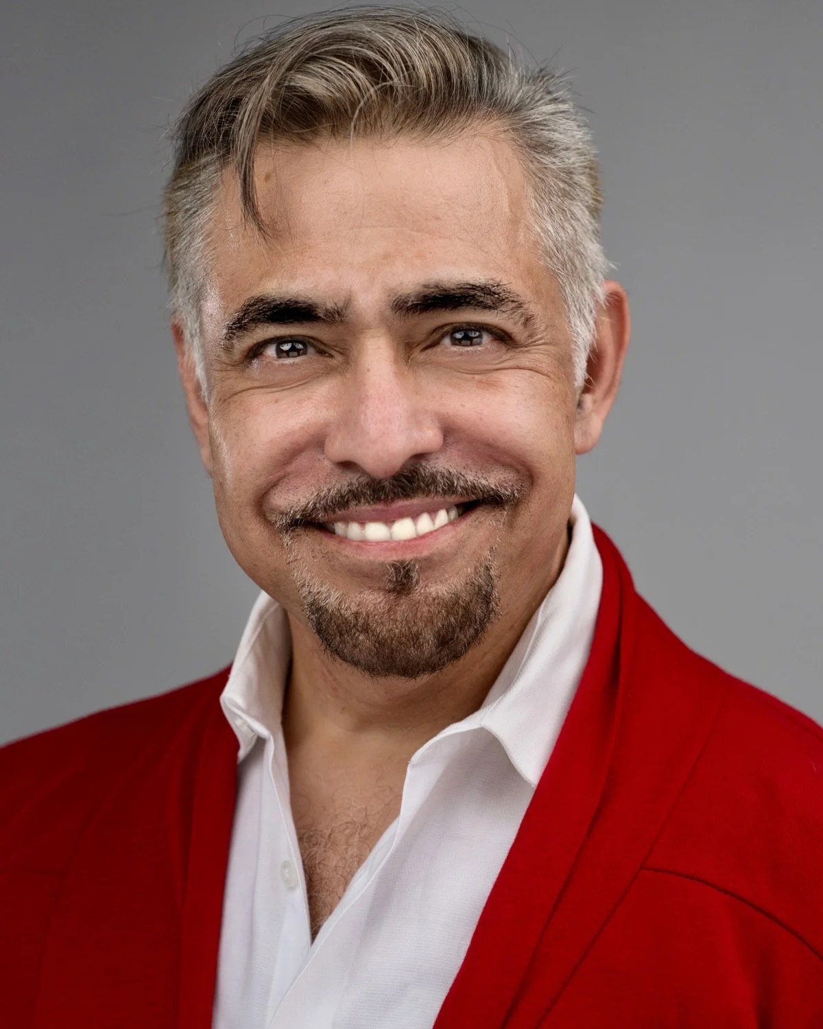 A middle-aged man with a goatee and smile wearing a white shirt and red blazer against a gray background.