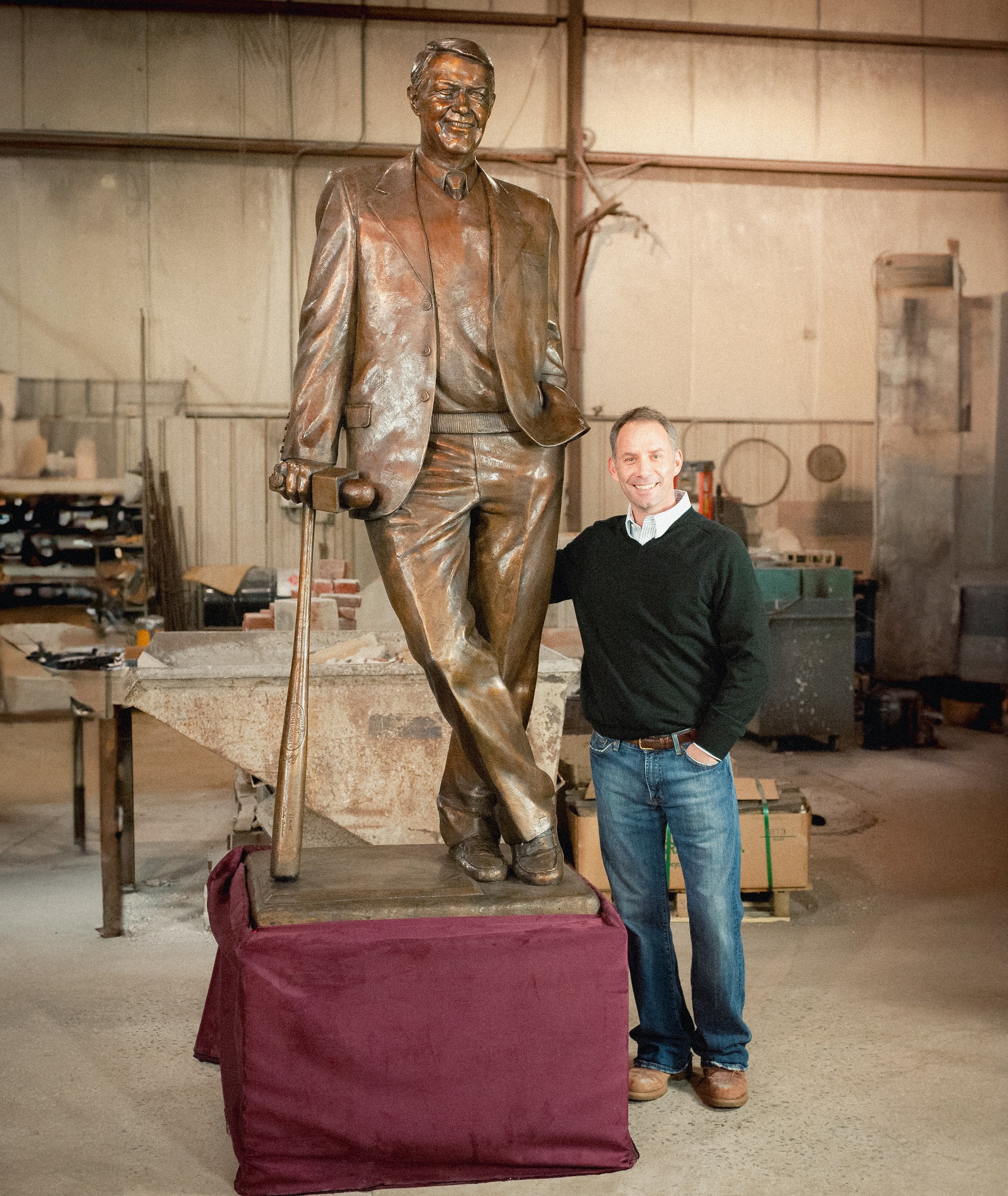 Man standing next to a large bronze statue of a man in a suit, holding a cane, in a workshop.