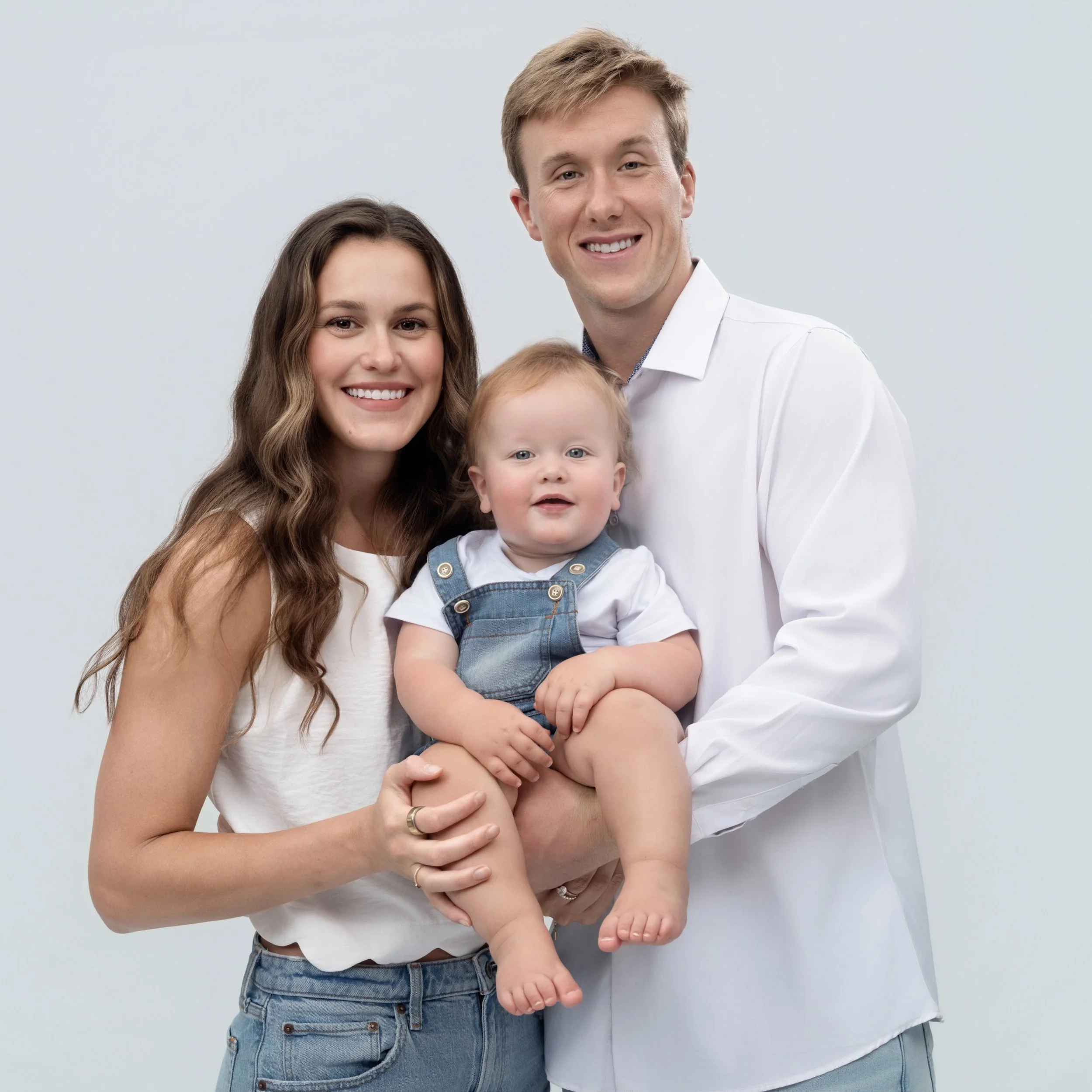 A smiling family of three with a woman, a man, and a baby, posing together against a plain light background.