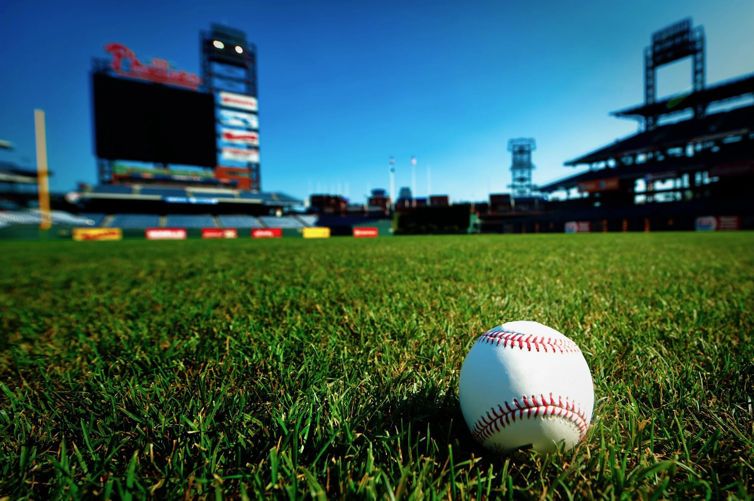 Close-up of a baseball on the grass baseball field with empty stadium seats and scoreboard in the background during daytime.