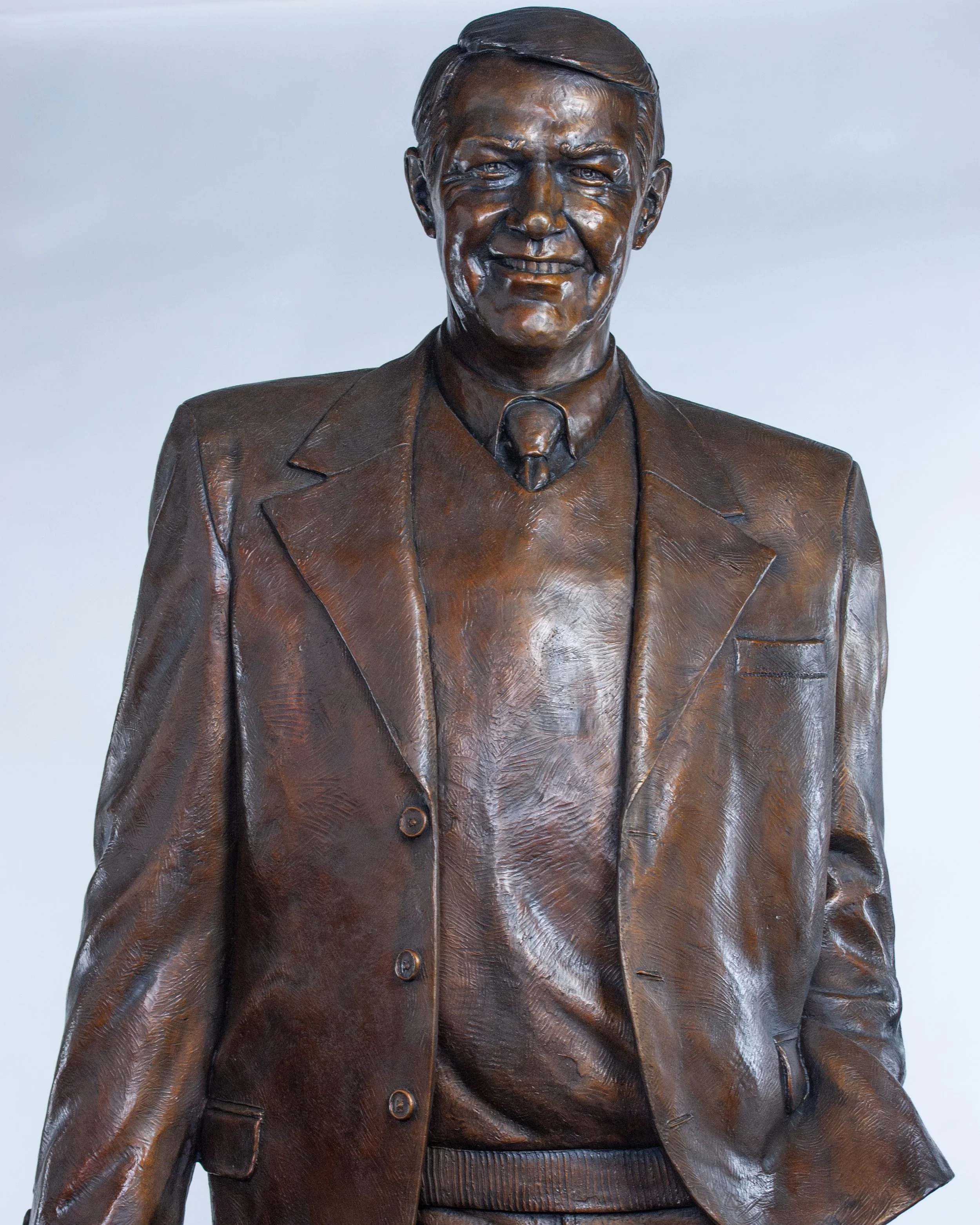 A bronze statue of a man in a suit with a tie, smiling, with a plain light background.