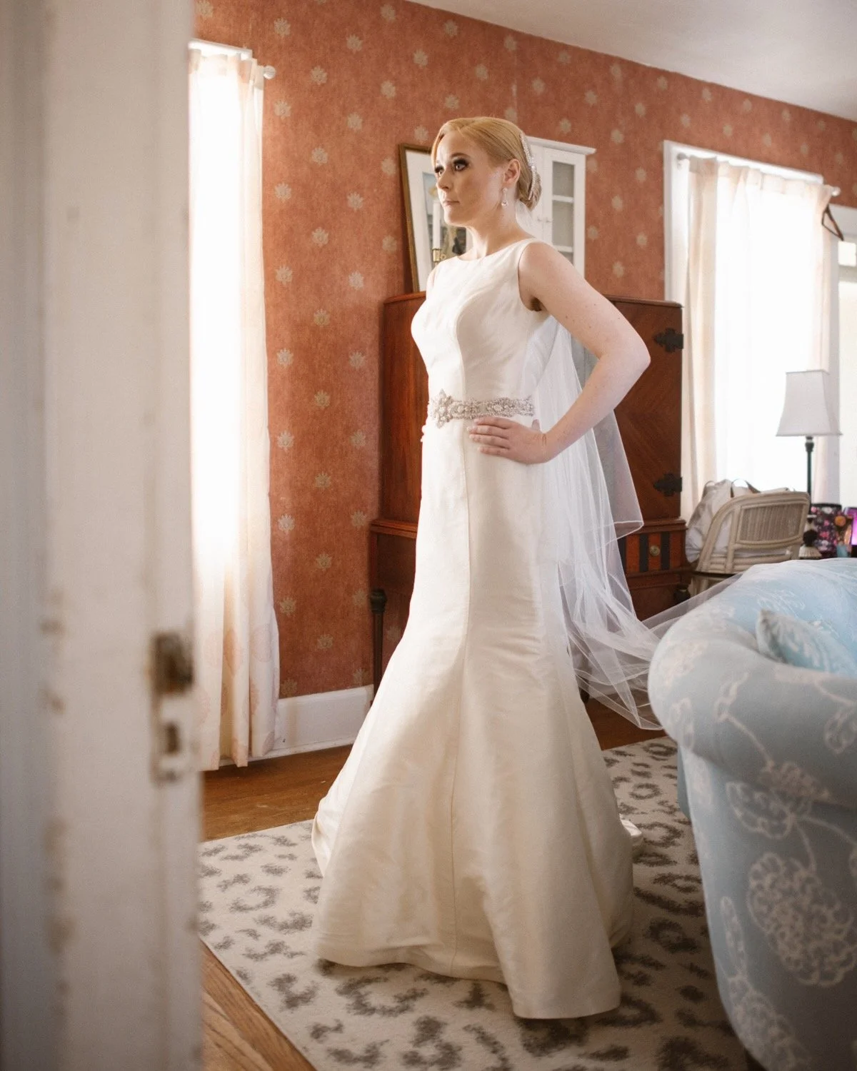 A bride in a white wedding gown standing in a bedroom with floral wallpaper, sunlight through windows, and a patterned rug.