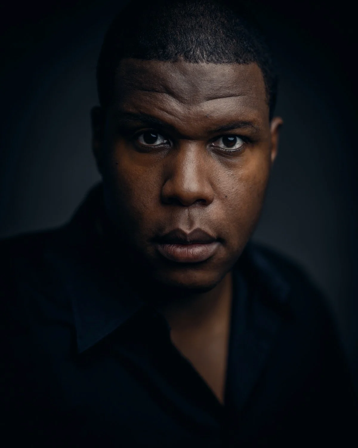 Close-up portrait of a young African American man with a serious expression, dark background, and black shirt.