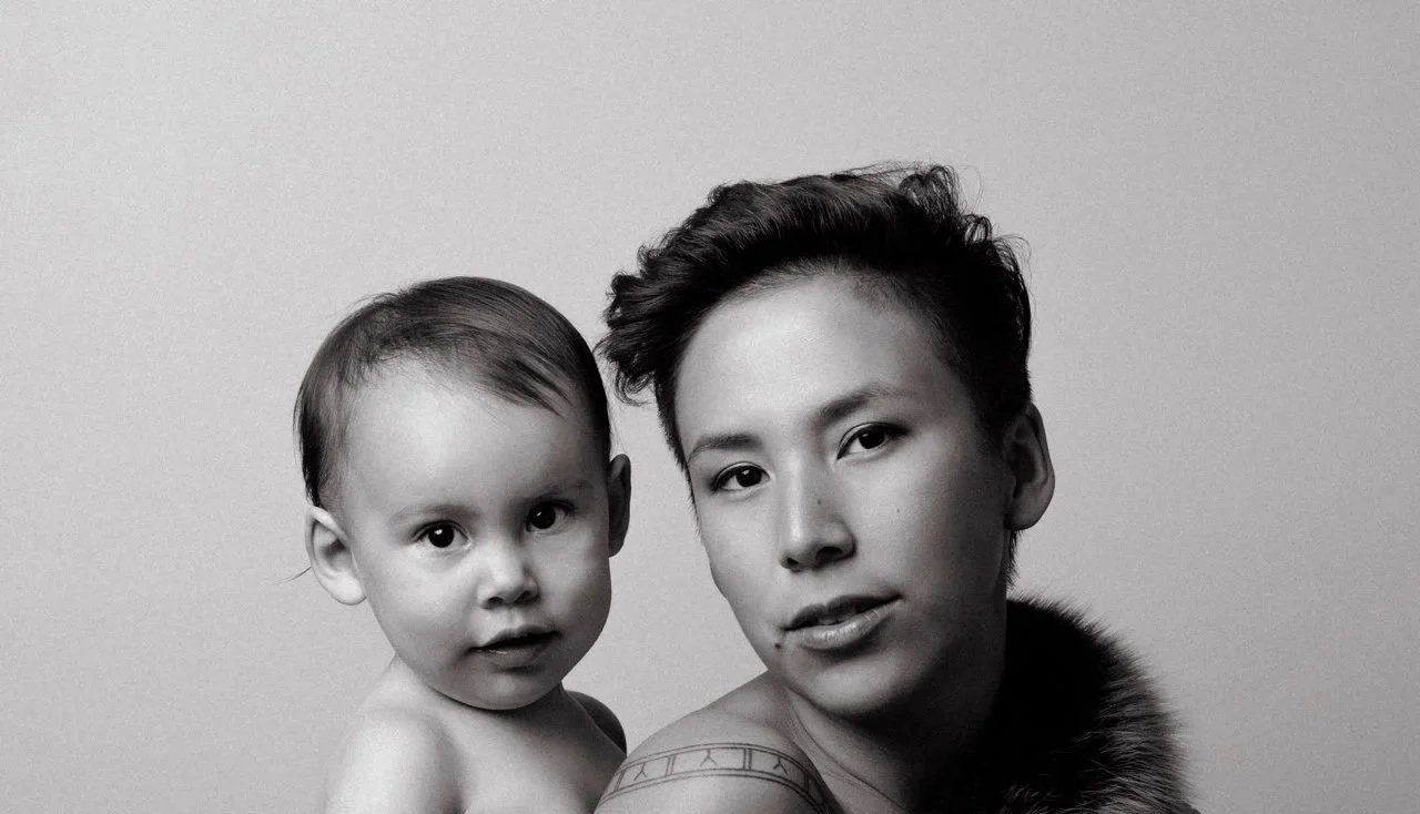 Black and white portrait of a young woman with short hair and a tattoo, holding a toddler with short hair, both looking at the camera against a plain background.