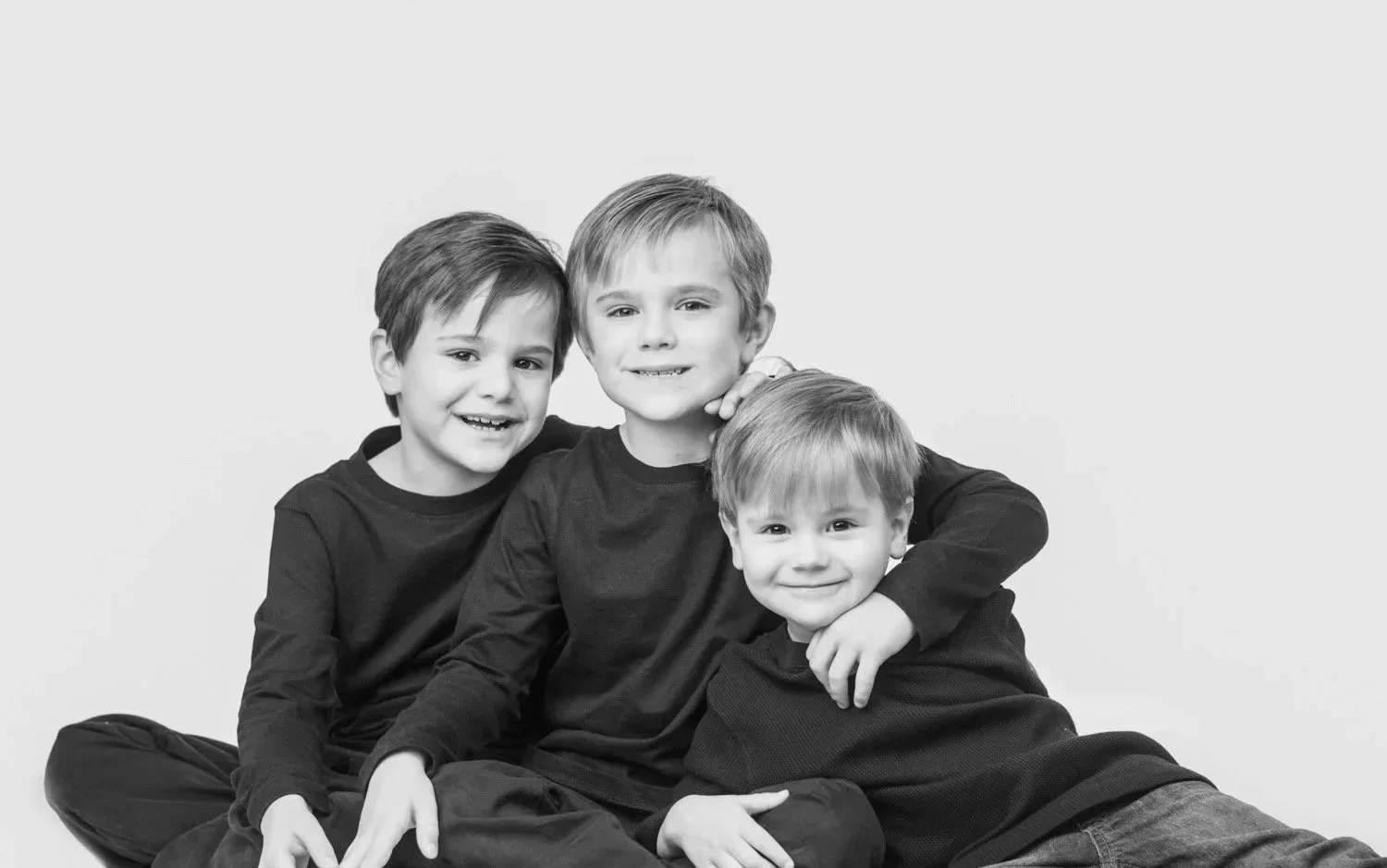 Three young boys sitting together, smiling, in a black-and-white photo.