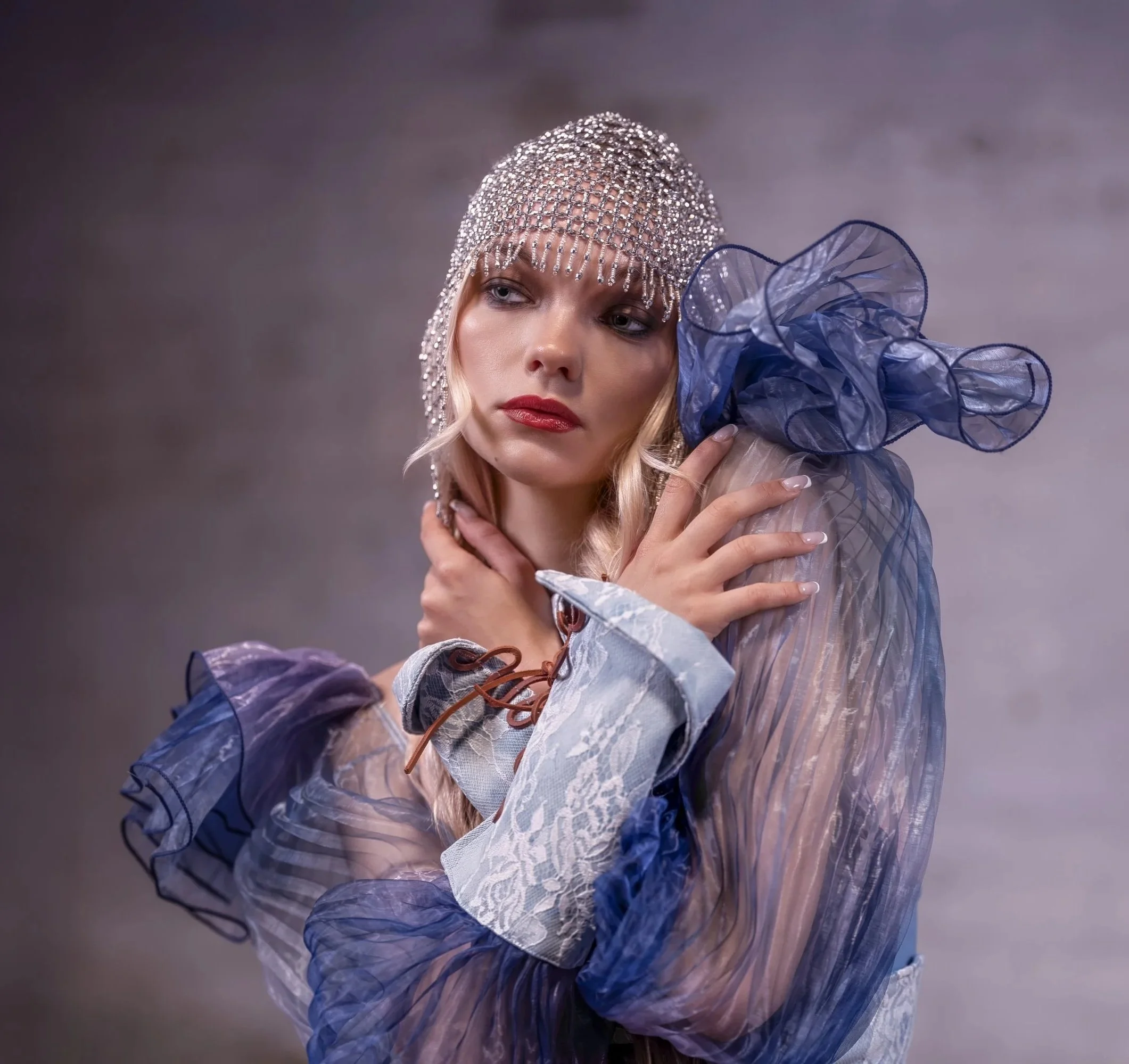 A woman wearing a beaded, silver headpiece and a sheer, blue, ruffled blouse with lace cuffs. She has fair skin, blonde hair, and red lipstick, and is touching her neck and shoulder with a pensive expression against a neutral, gray background.