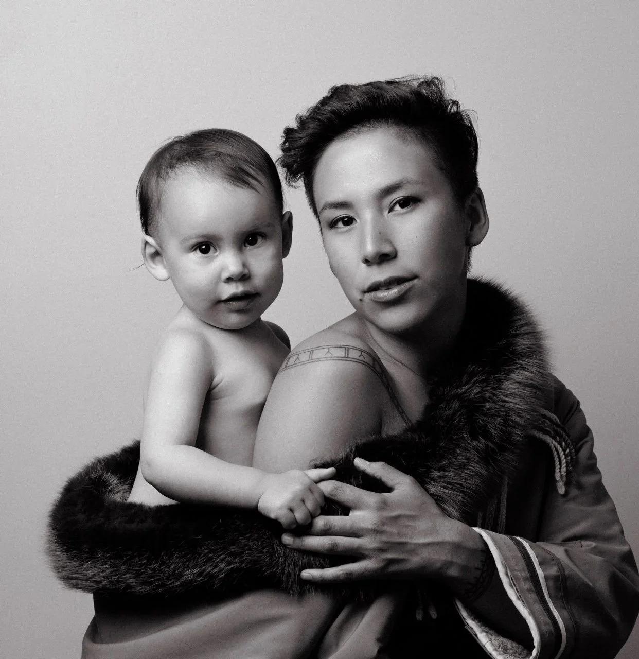A black and white photo of a woman with short hair and a young child, both looking at the camera. The woman is partially draped in a fur-lined coat, and the child's hand rests on her shoulder.
