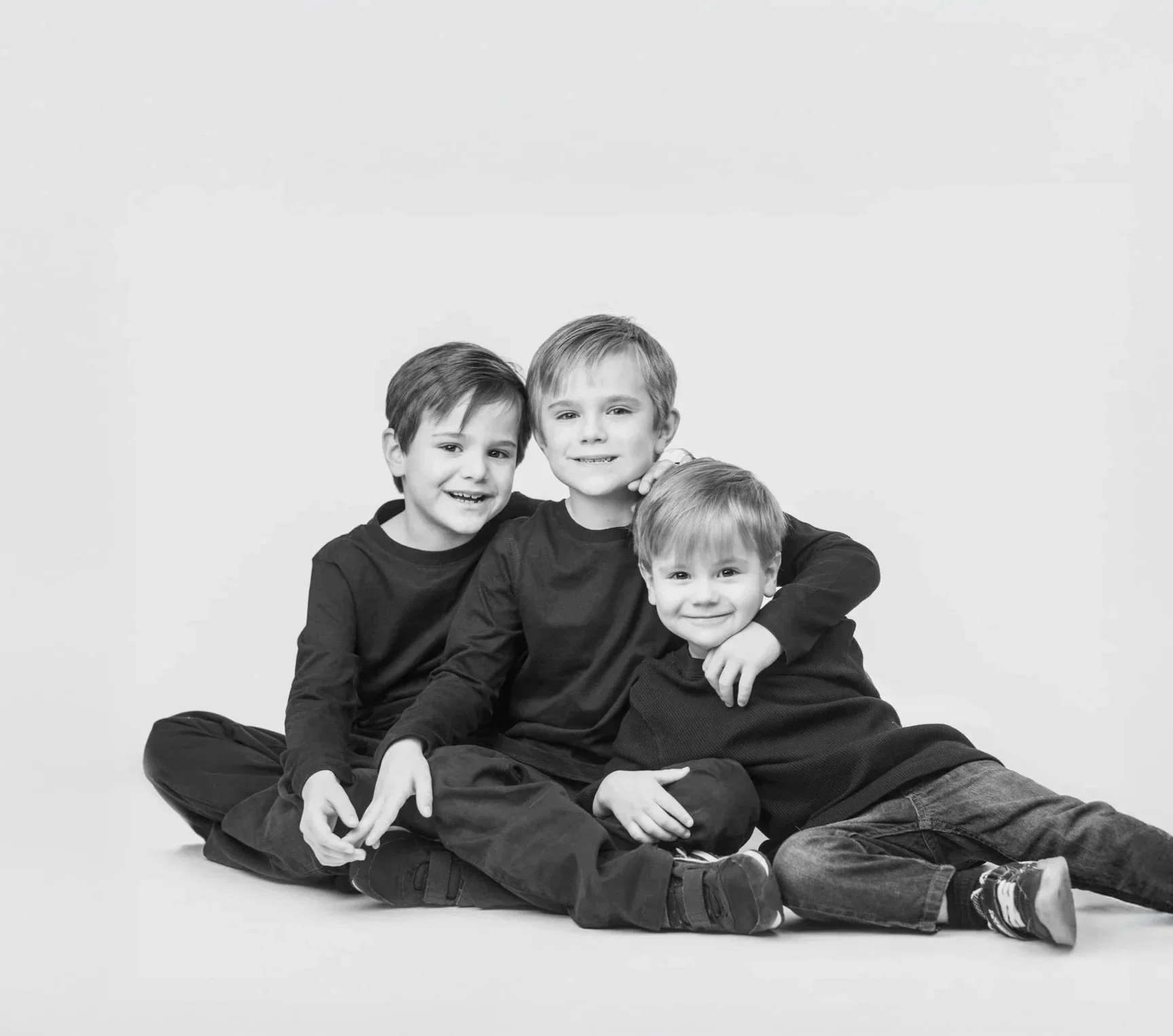Three young boys sitting on the floor, smiling, embracing each other in a black-and-white photo.