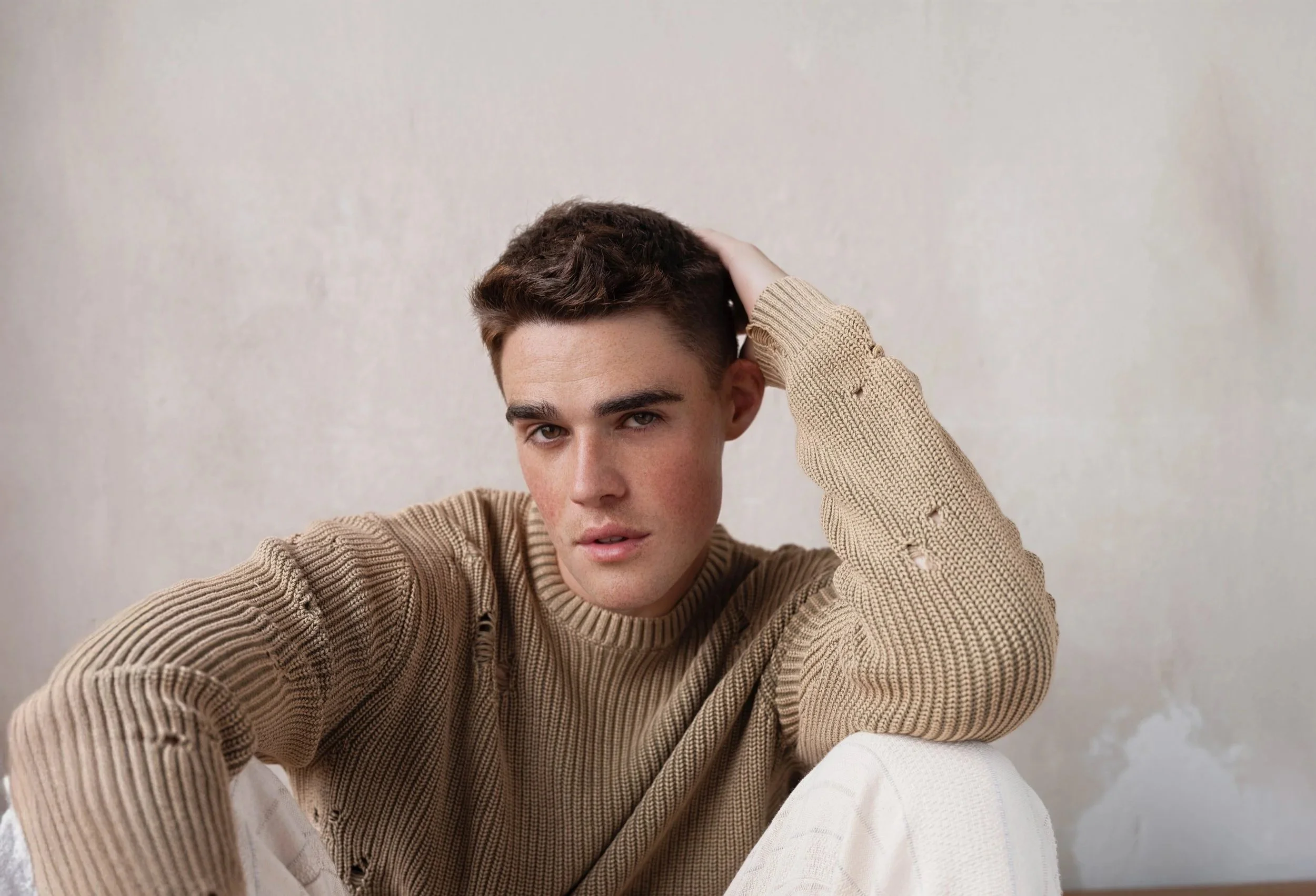 A young man with short brown hair, wearing a beige ribbed knit sweater with distressed details and white pants, posing indoors against a plain light-colored wall.